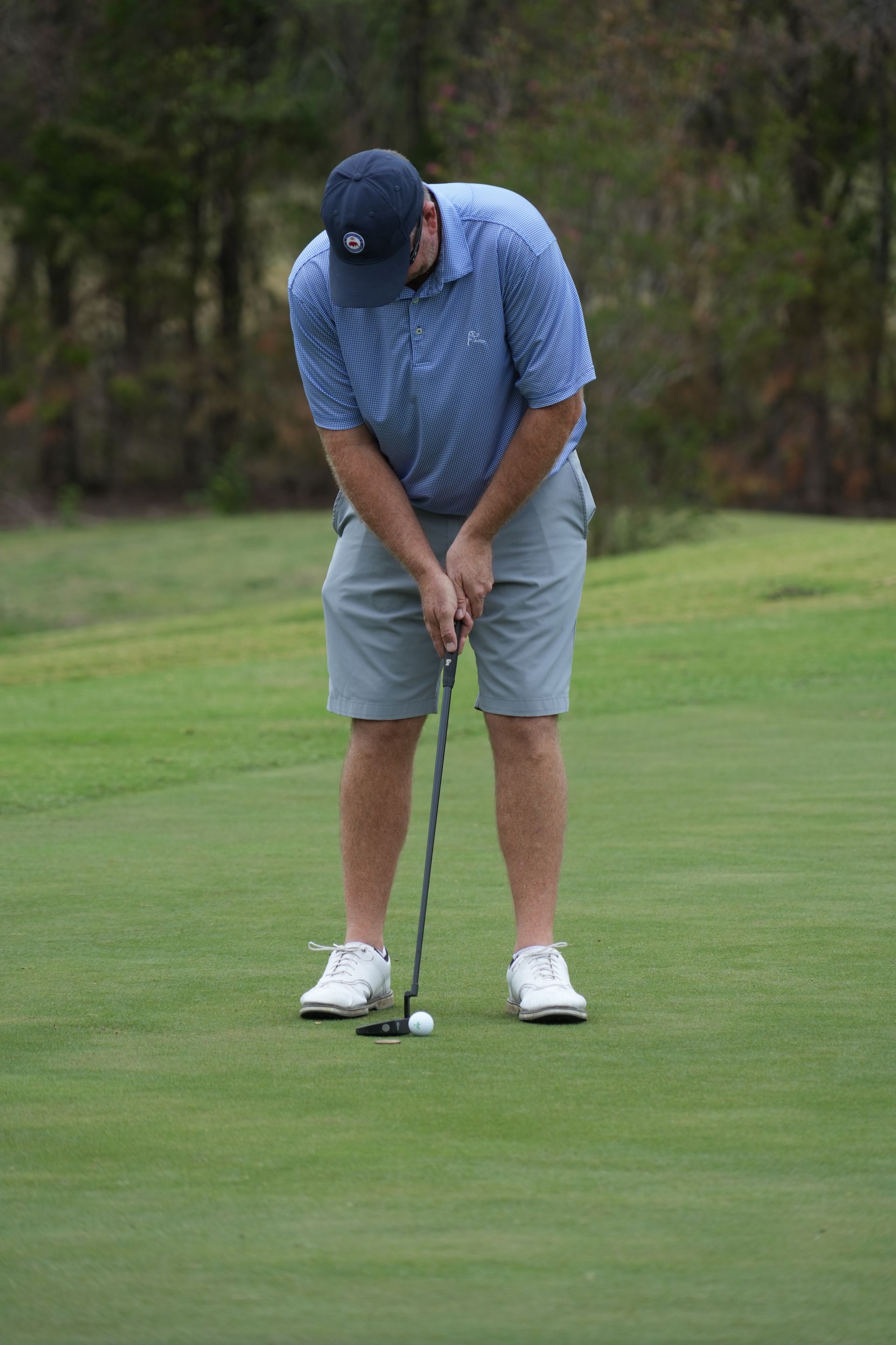 Golfer putting on green, wearing blue patterned shirt, gray shorts, and white shoes; ball in front of putter.