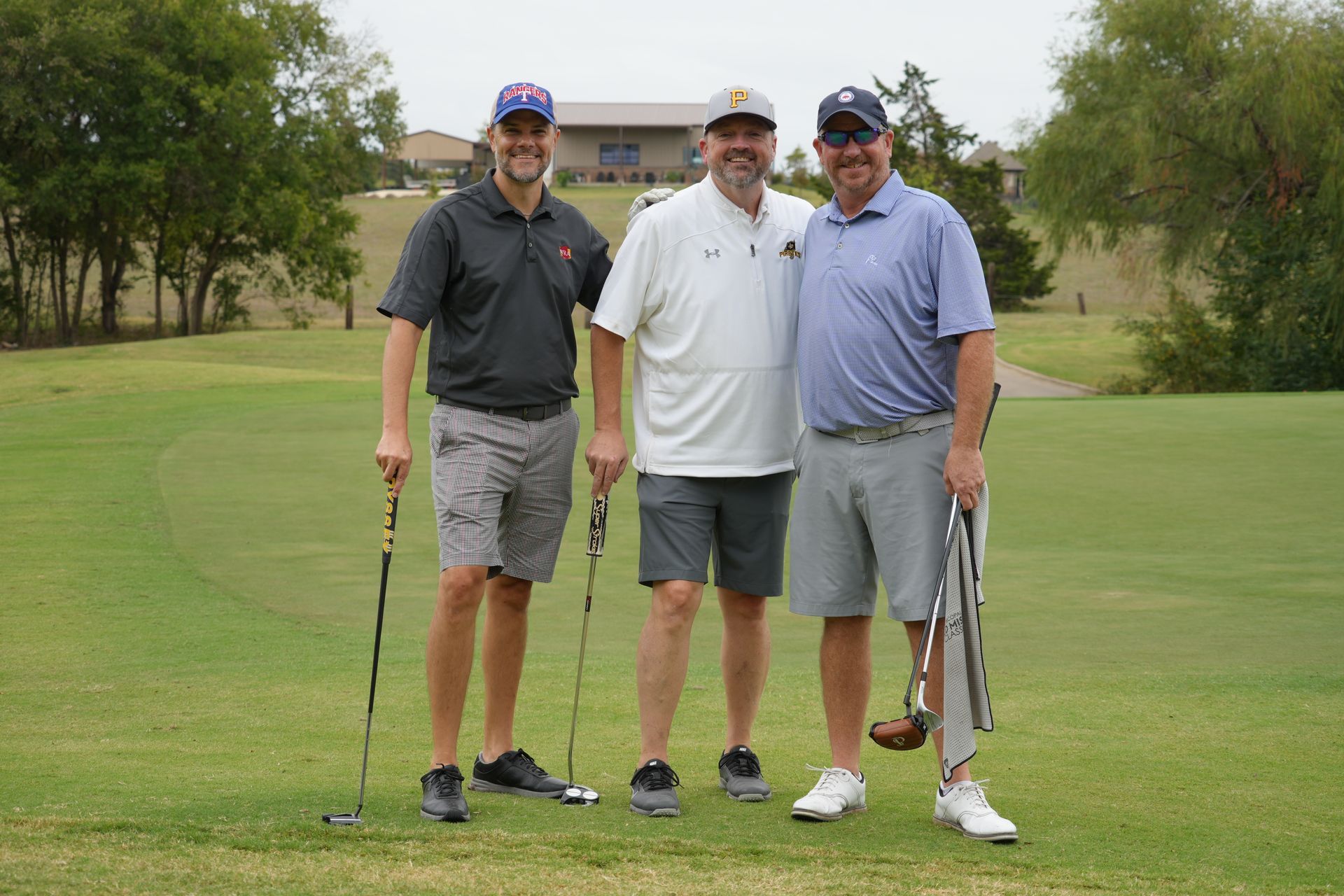Three men smiling on a golf course, holding clubs, in shorts and polo shirts.