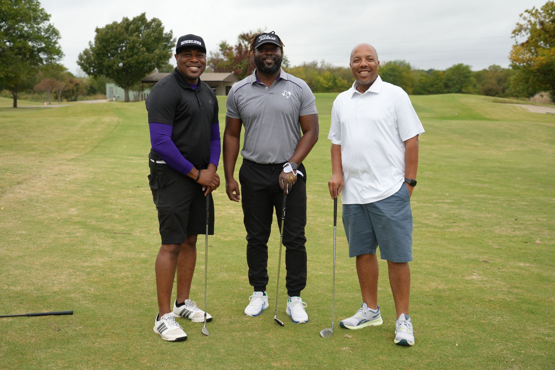 Three men on a golf course: posing with clubs, one purple arm sleeve, casual attire, smiling.