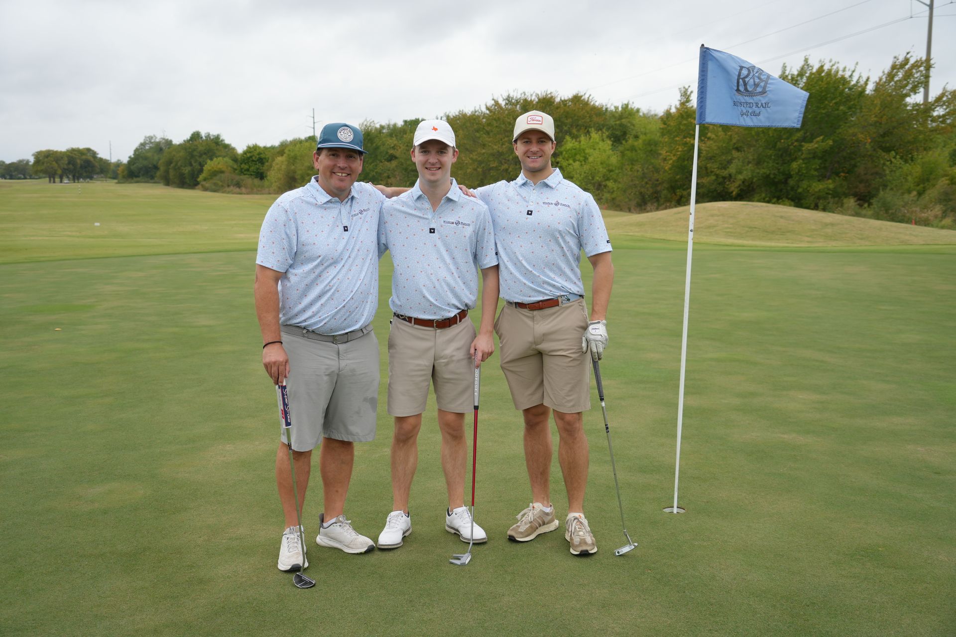 Three golfers pose on a green, wearing light blue shirts and khaki shorts, a blue flag in the background.