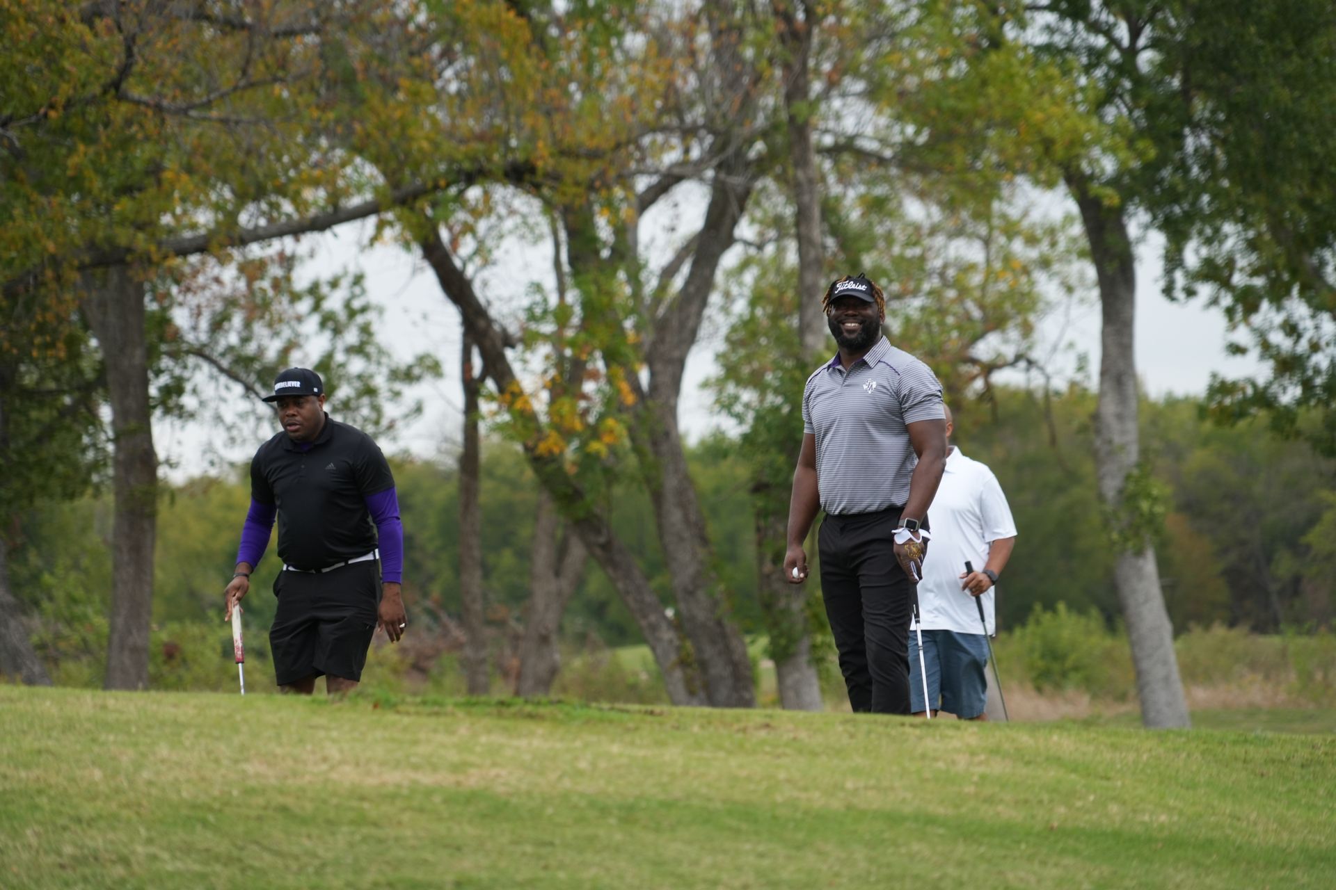 Two men playing golf on a grassy course under trees; one wears black and purple, the other gray and black.