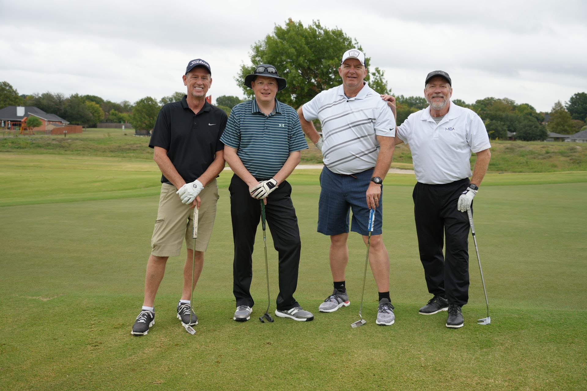 Four men on a golf course posing with clubs. They wear golf attire. Green grass and overcast sky in the background.