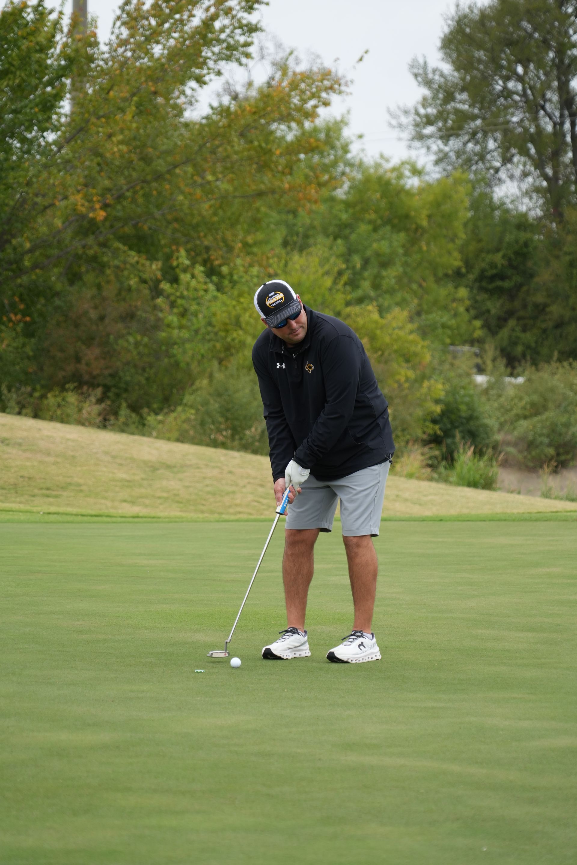 Man putting a golf ball on a green, wearing black jacket, grey shorts, and white shoes. Outdoors.