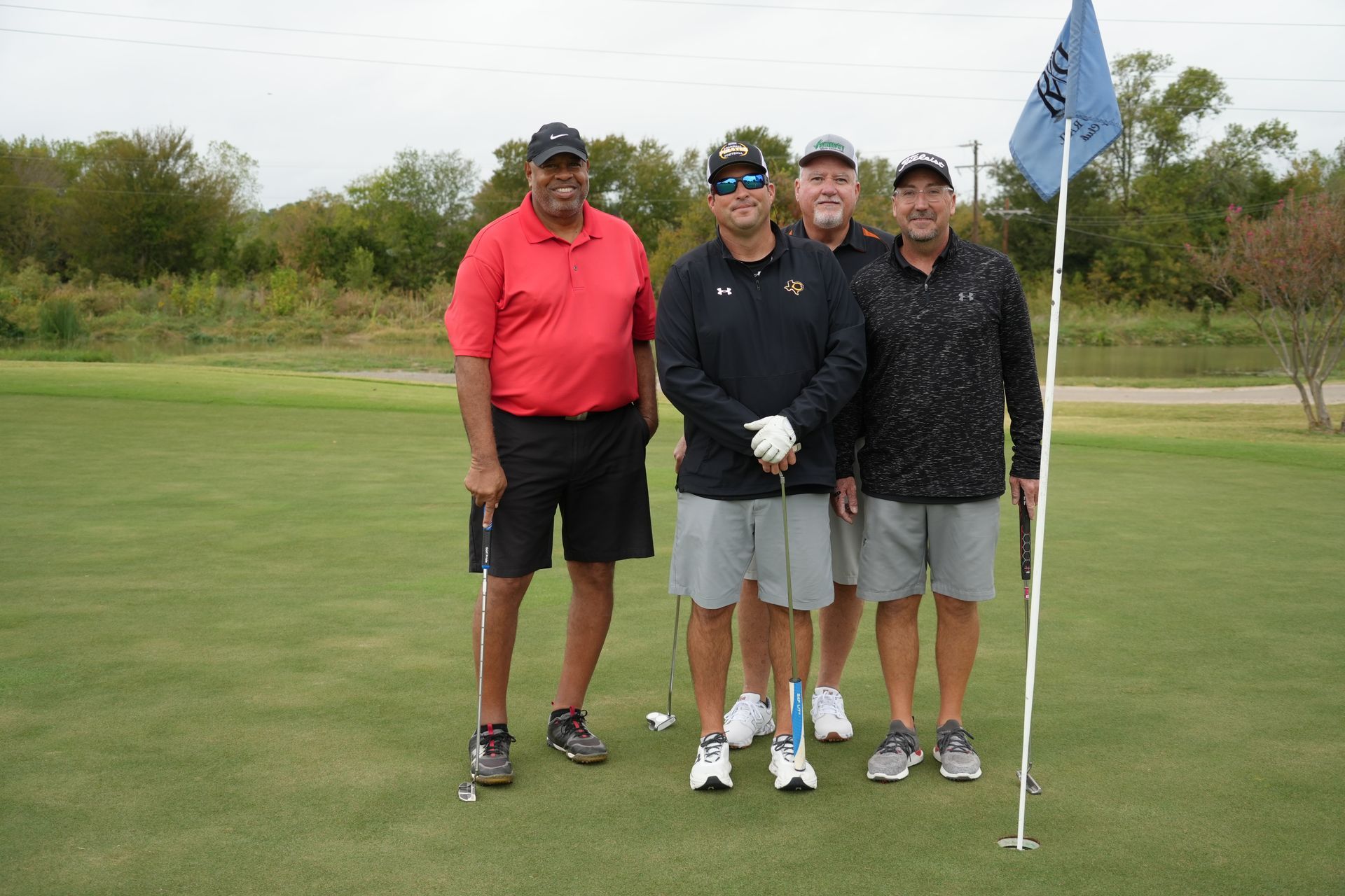 Four men on a golf course, smiling, posing near a flag. One man holds a golf club.