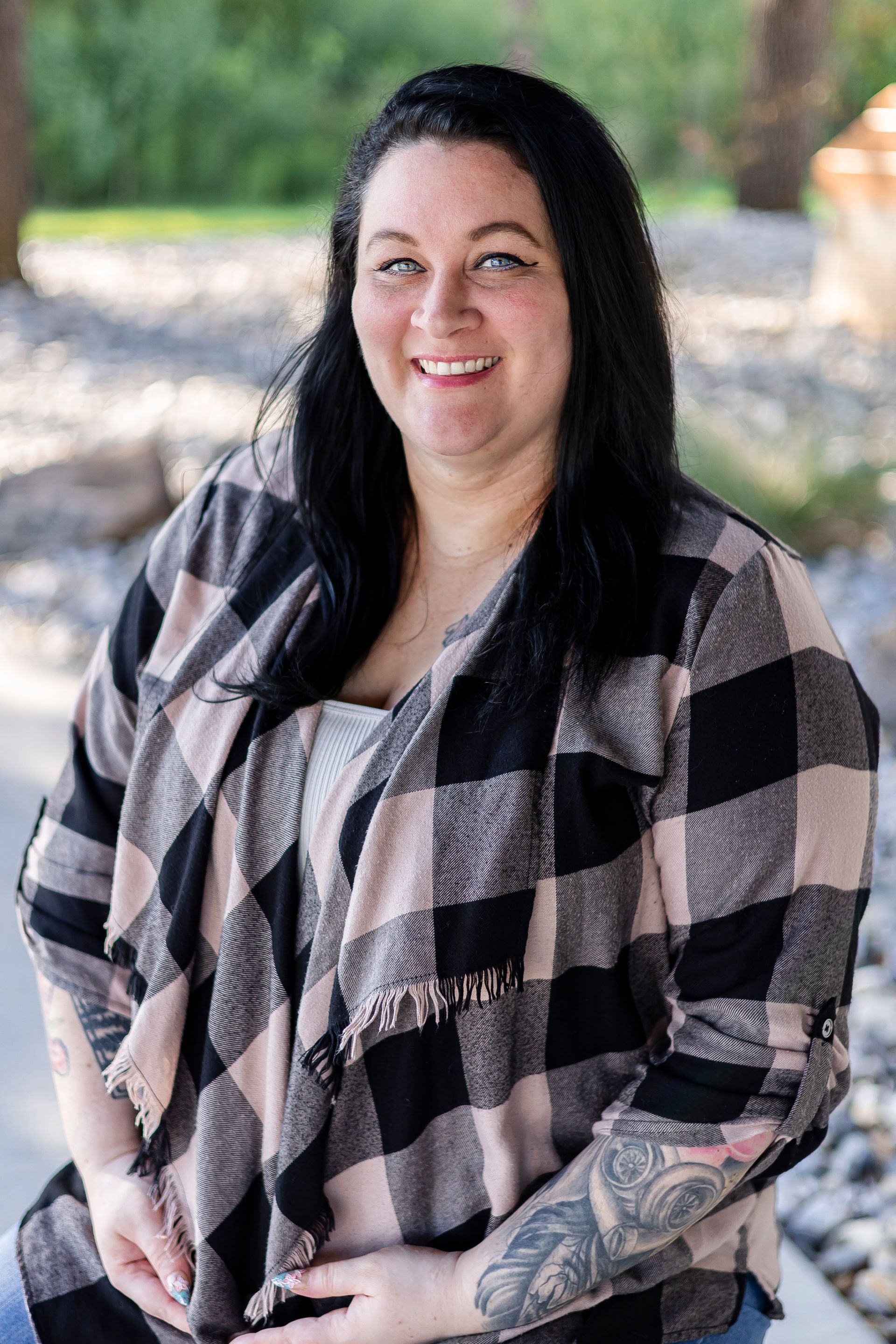 Woman with dark hair smiling, wearing black and pink plaid cardigan. Outdoors, sunny.