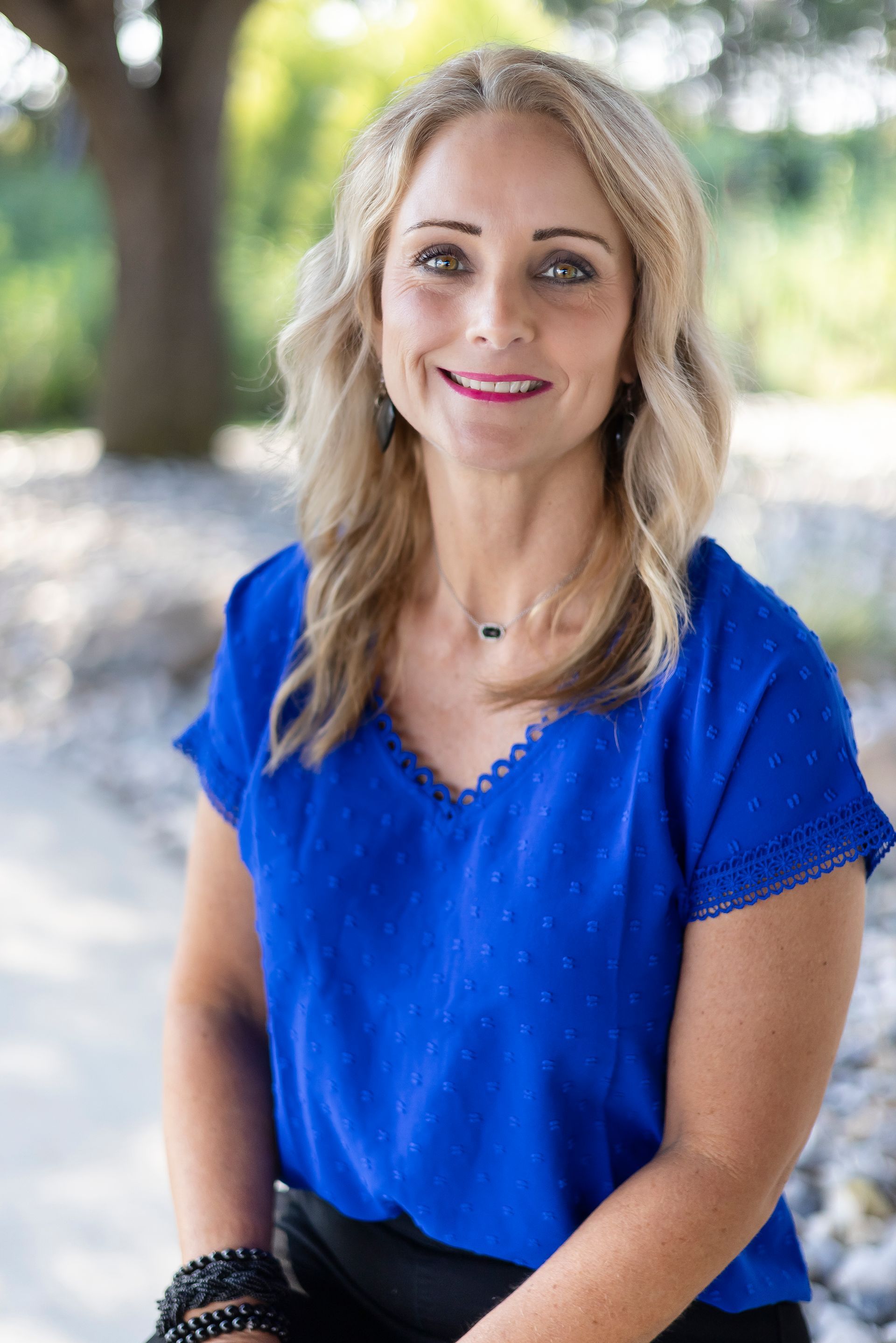 Blonde woman in blue shirt smiles outdoors, sitting with arms crossed, next to tree.