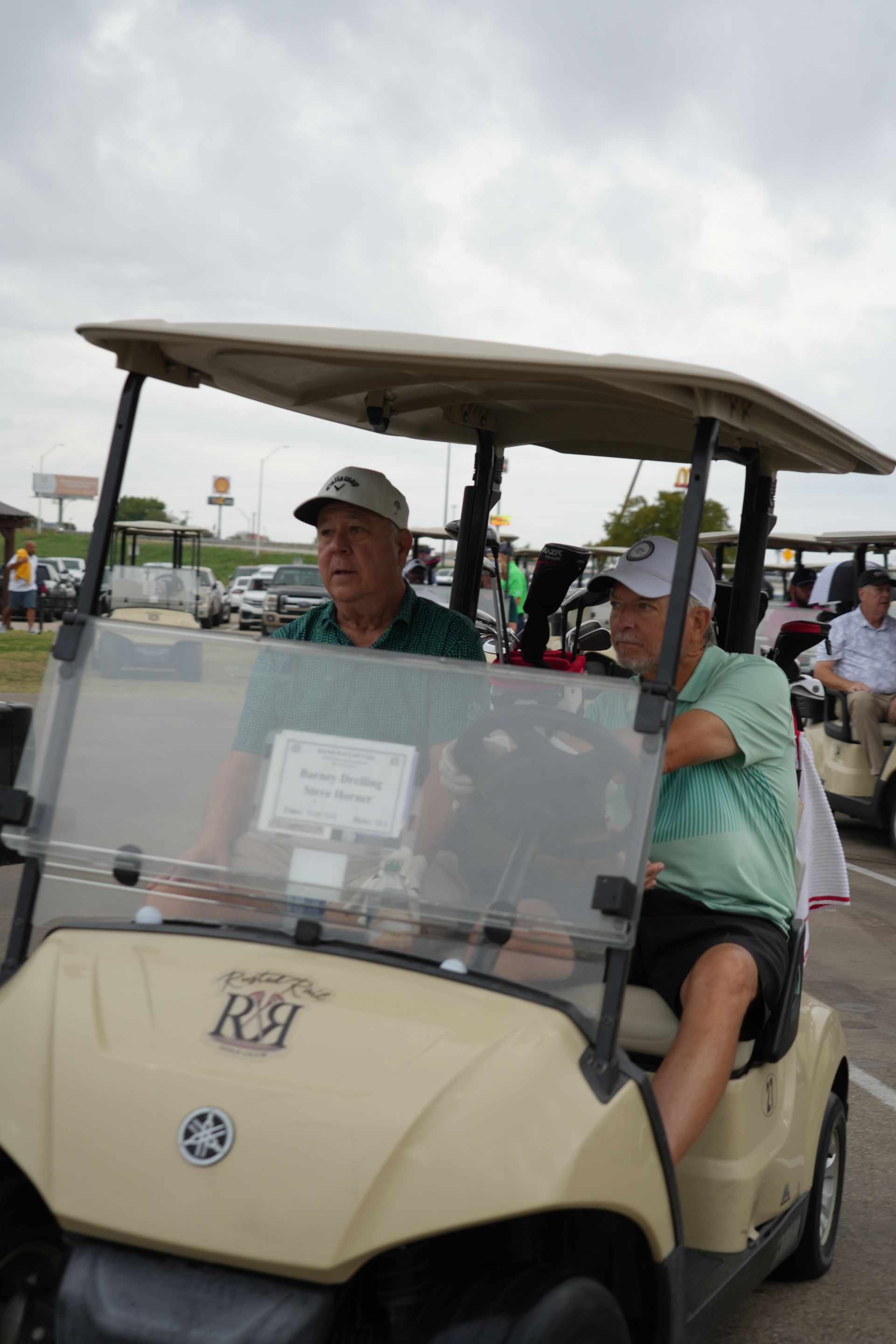 Two men in golf cart, beige cart, driving on a cloudy day.