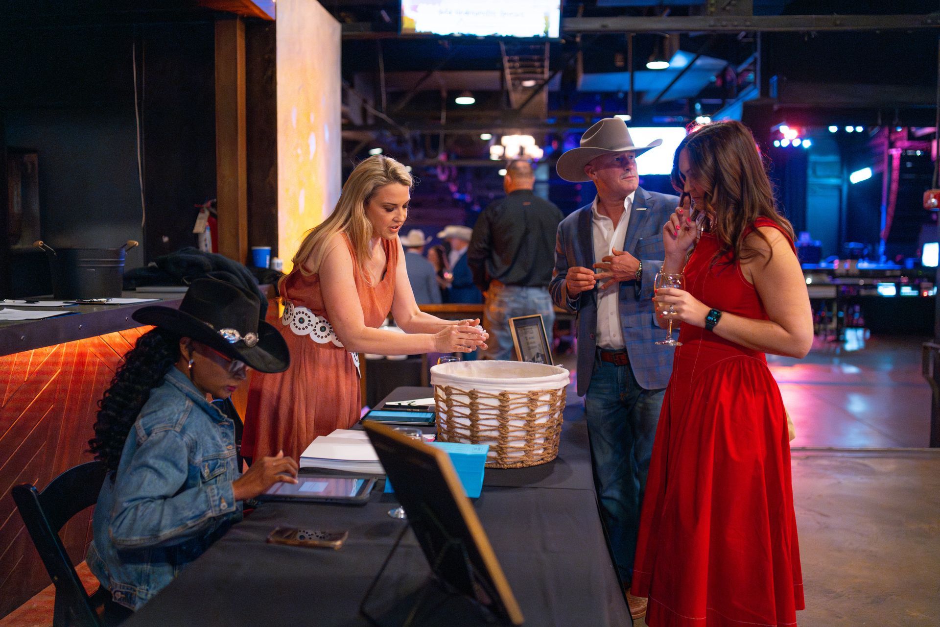 People at an event, some around a table. Woman in red dress talks with another, while a woman in a witch hat works the table.