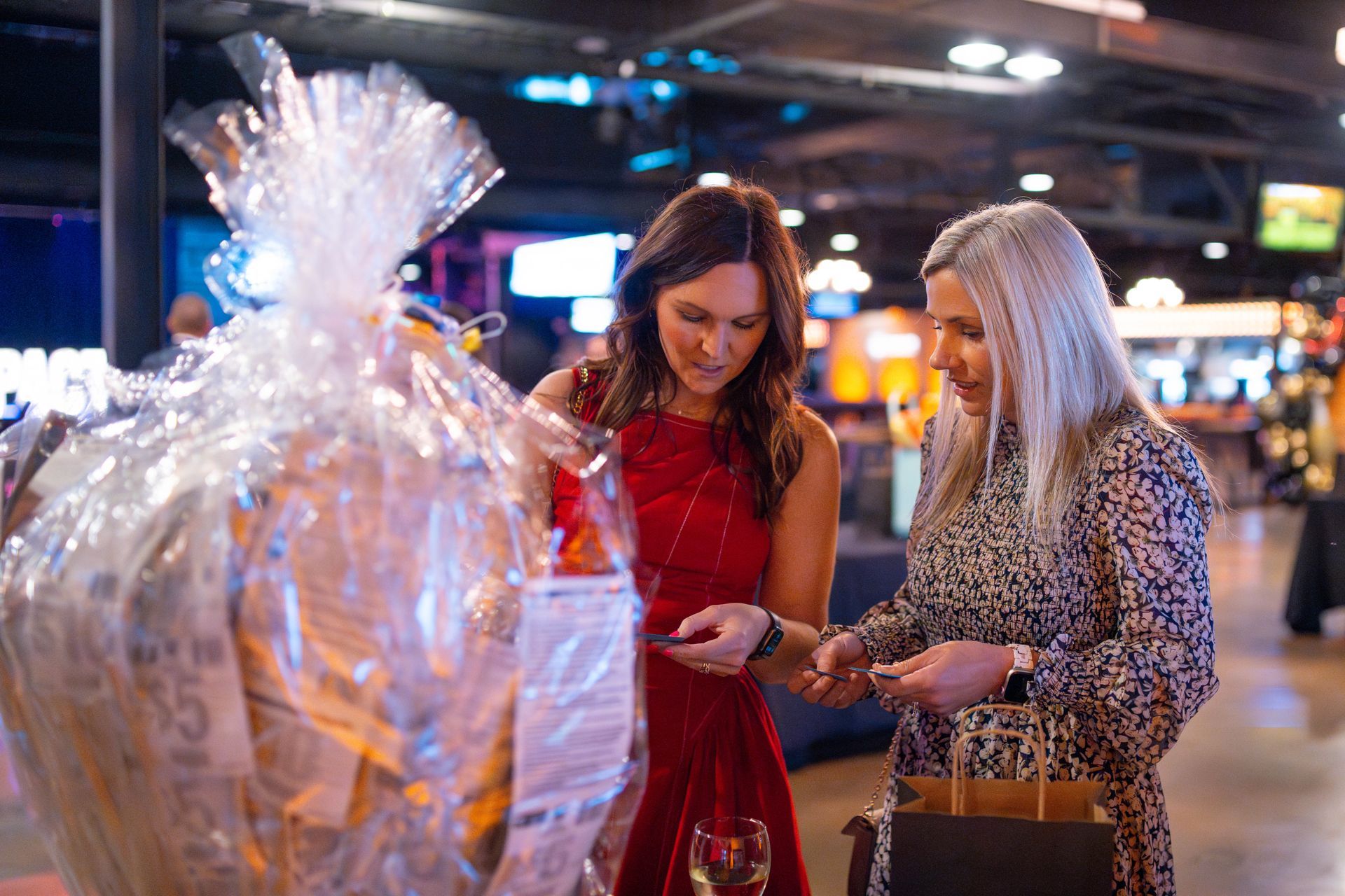 Two women examining a gift basket in a festive indoor setting. One wears a red dress, the other a patterned blouse.