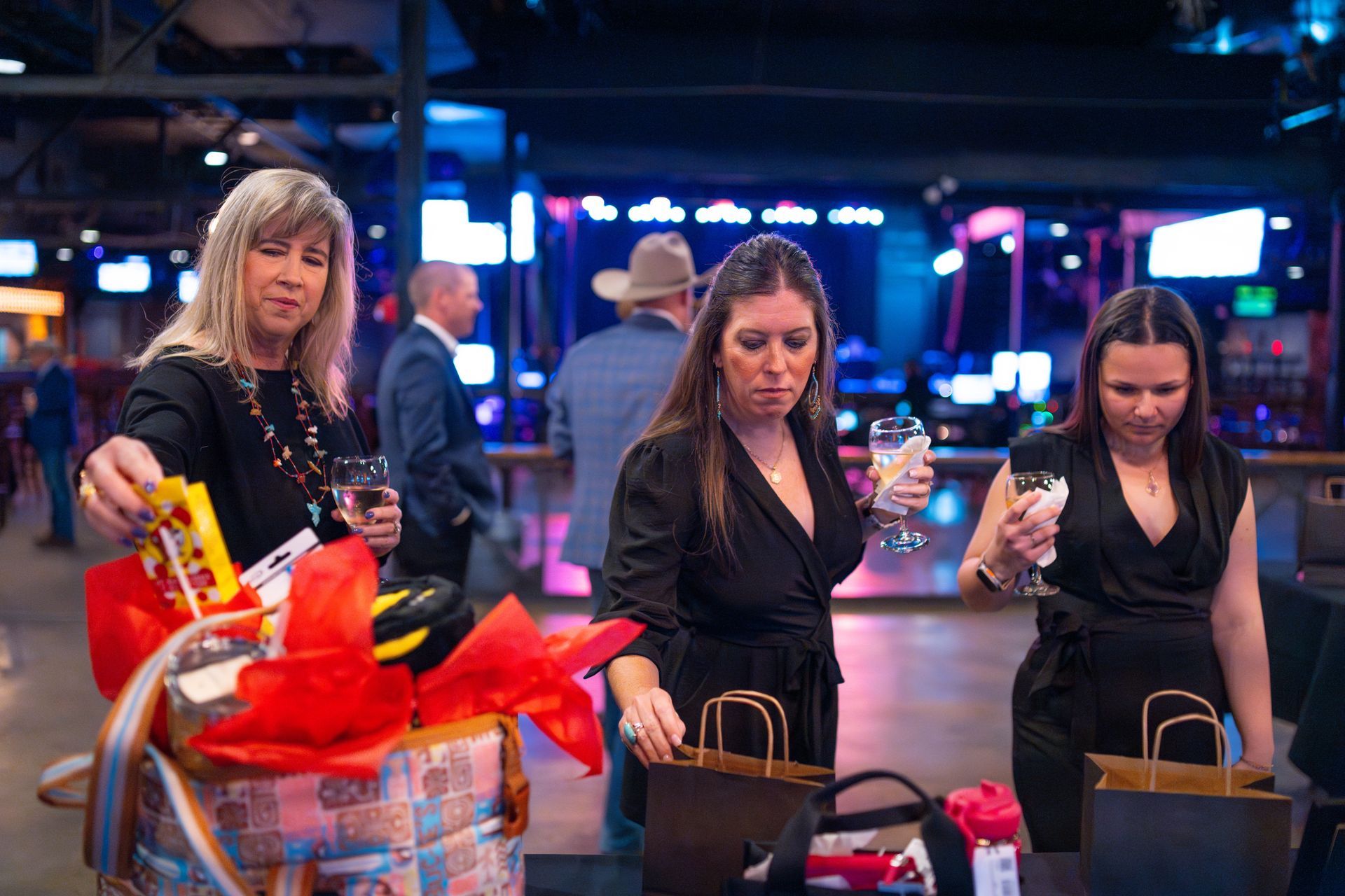 Three women at a party with gifts. One holds a gift basket, others look at bags. Indoor event, low lighting.