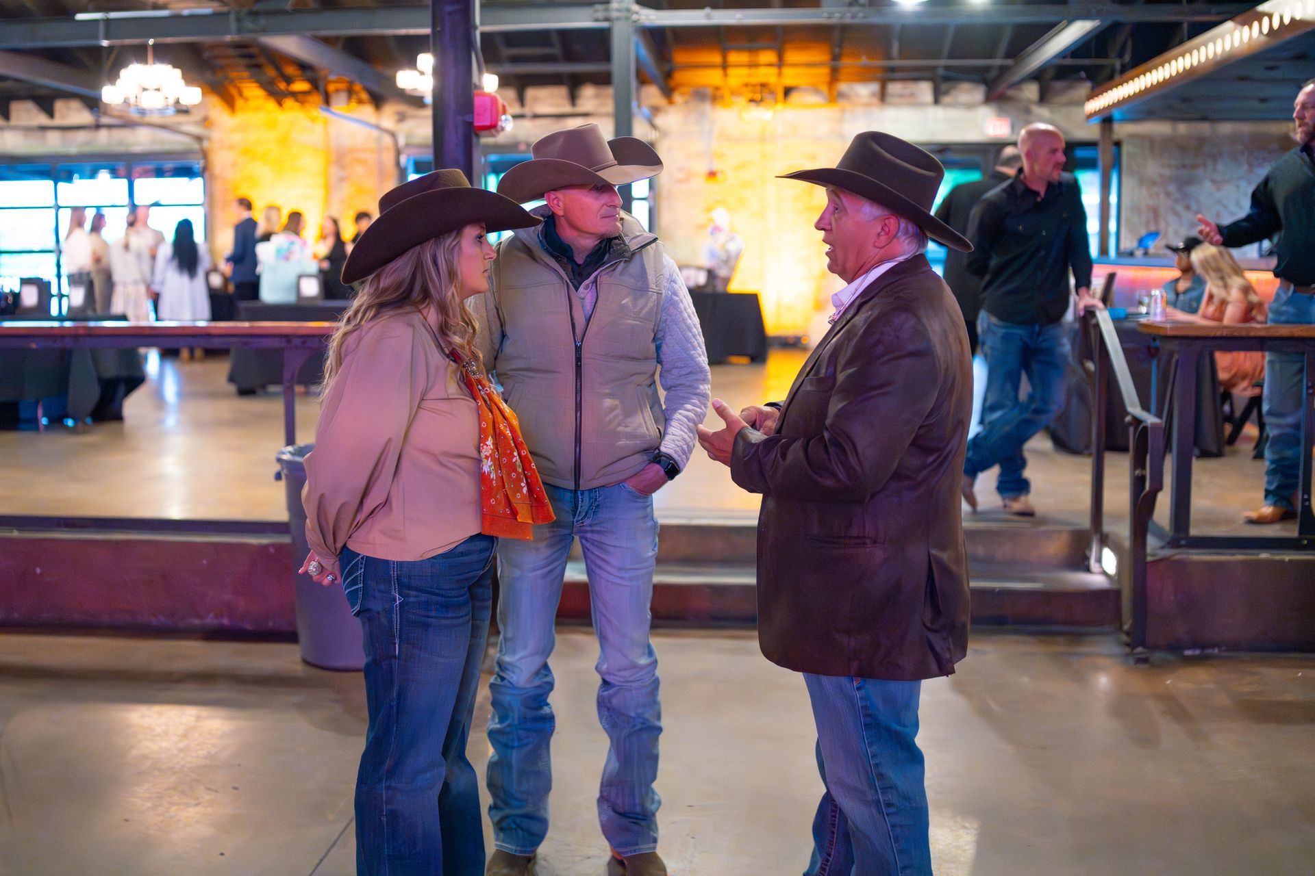 Three people in cowboy hats converse indoors; a woman, a man in a vest, and a man in a coat.