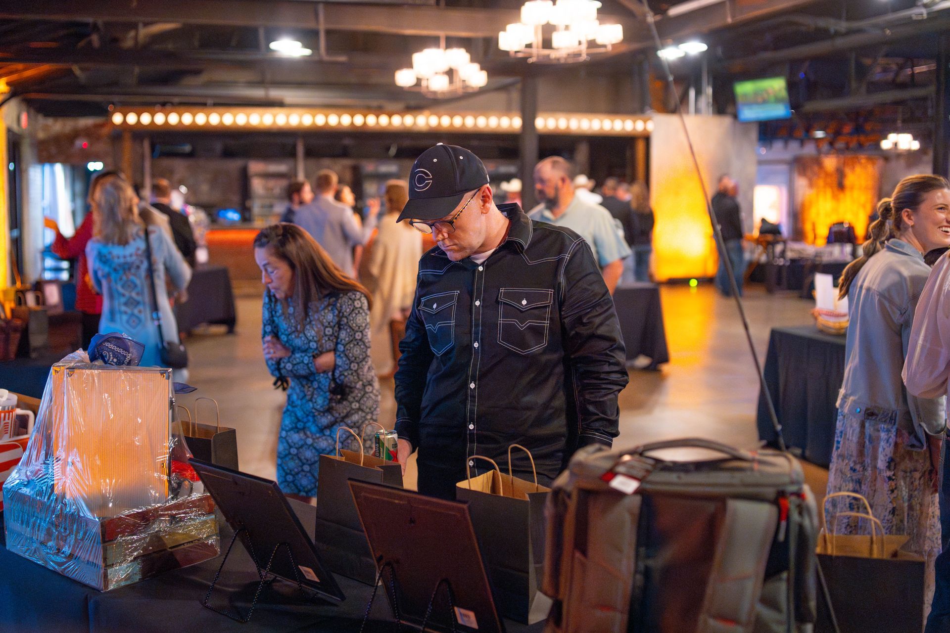 People browse items at an event. A man in a black shirt looks at table display with a girl. Venue is dimly lit.