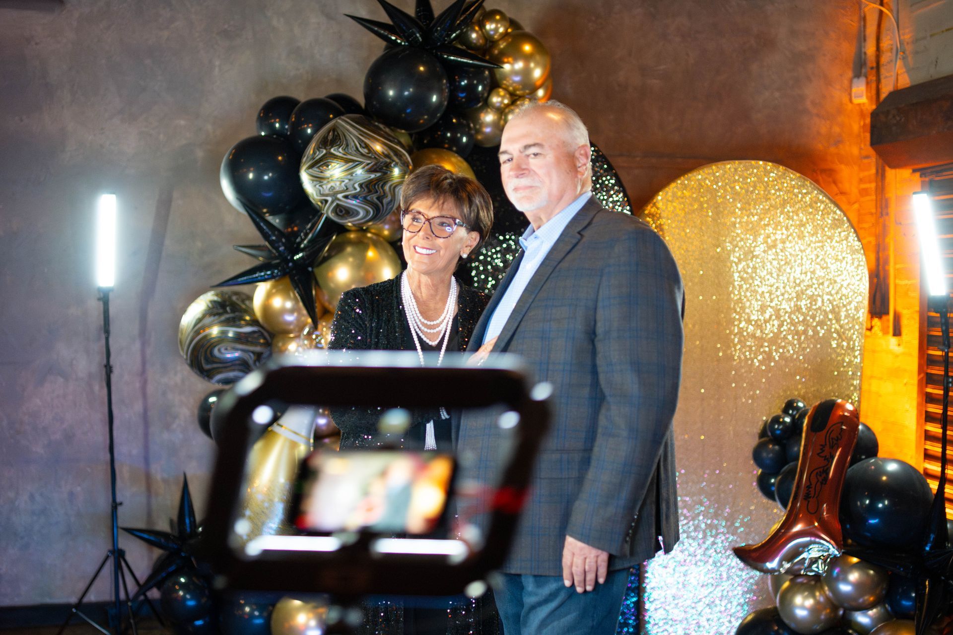 Couple poses for photo; black and gold balloon backdrop, illuminated photo booth setup.