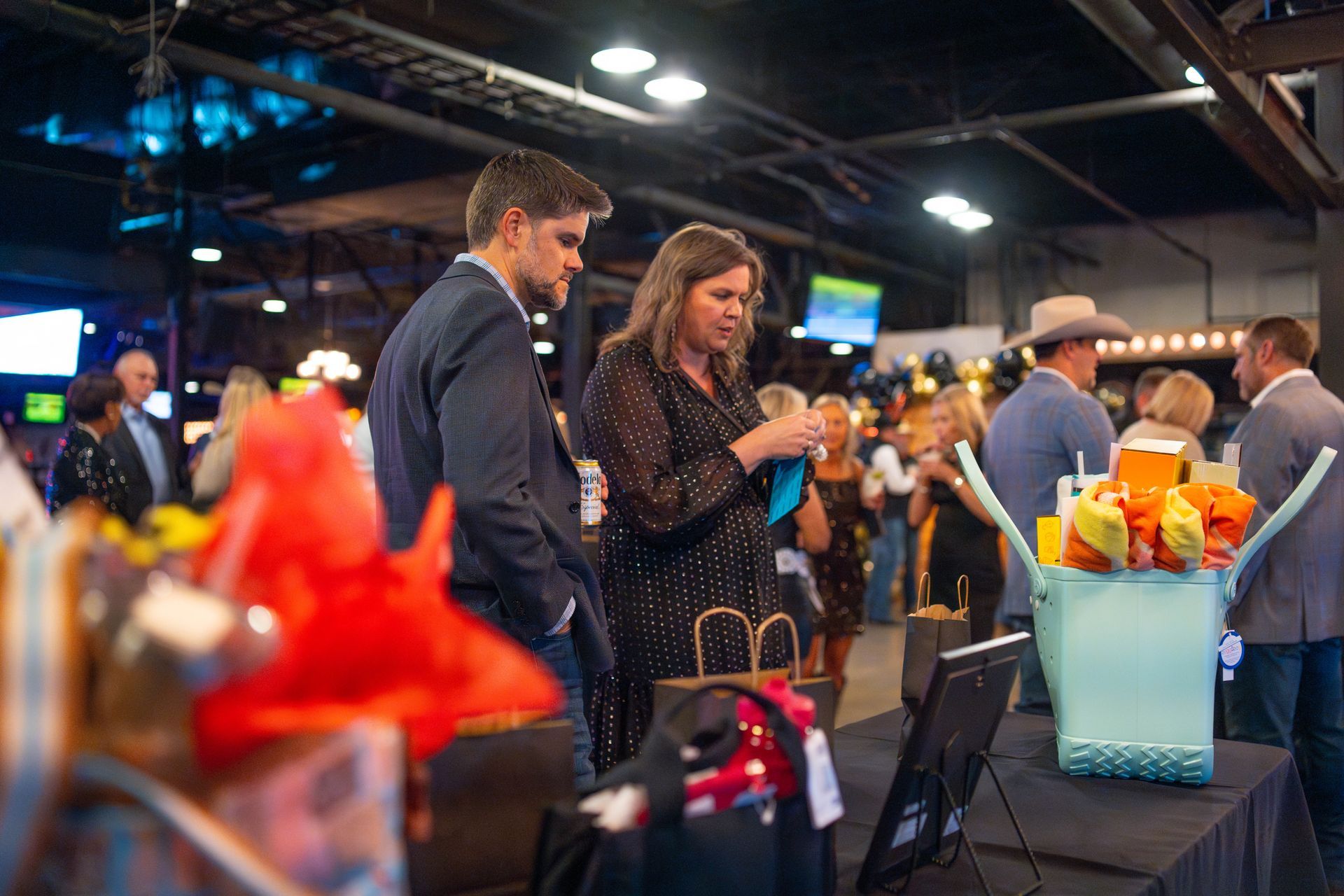 Two people at a gala examining gift bags. Others mingle in a large hall.