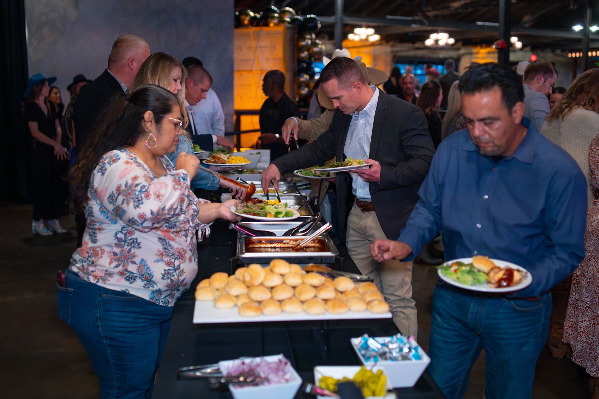 People at a buffet, serving themselves food. Rolls, salads, and other dishes are displayed on the table.