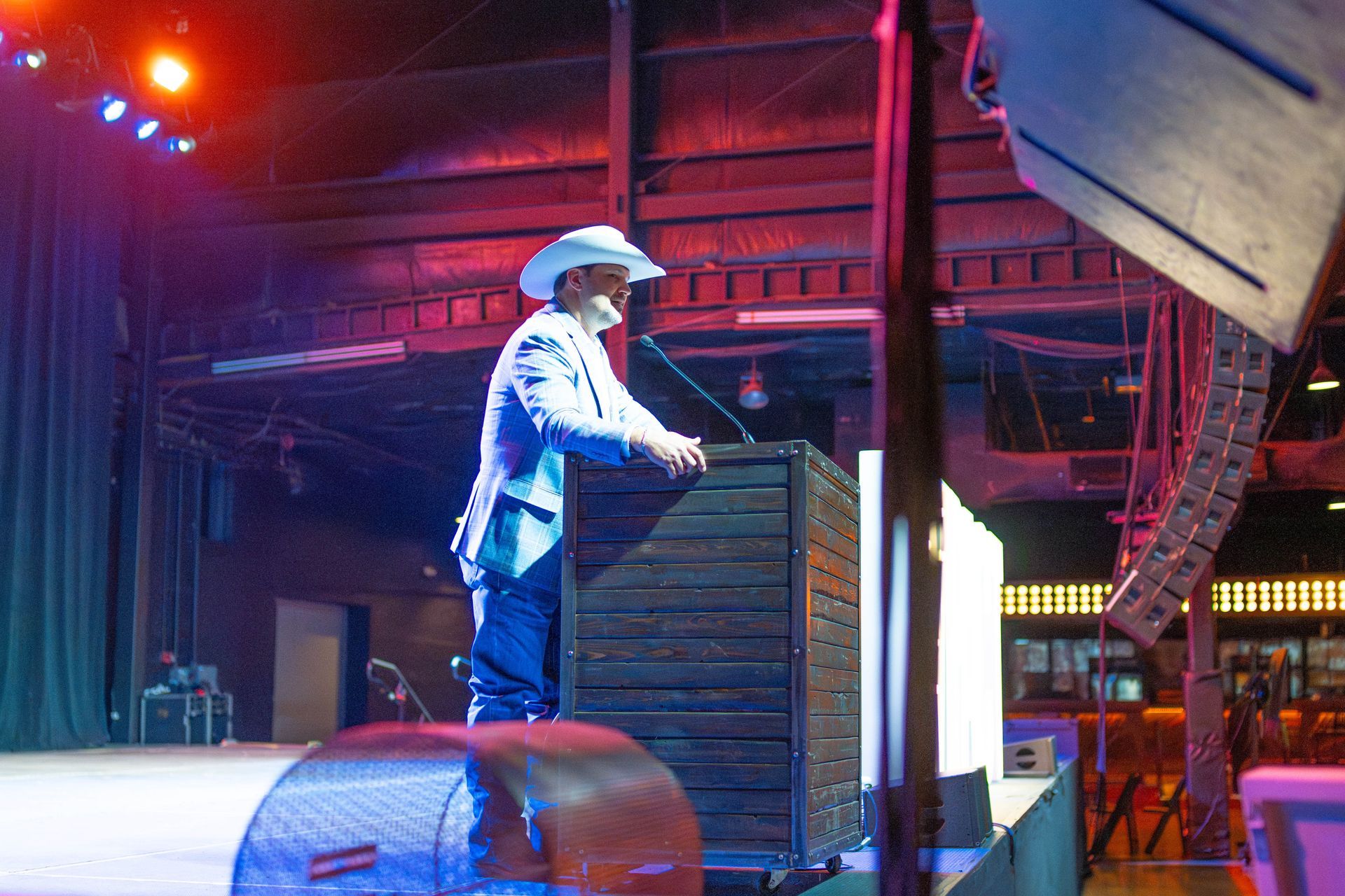Man in a cowboy hat speaking at a podium on a stage, lit by blue and red lights.