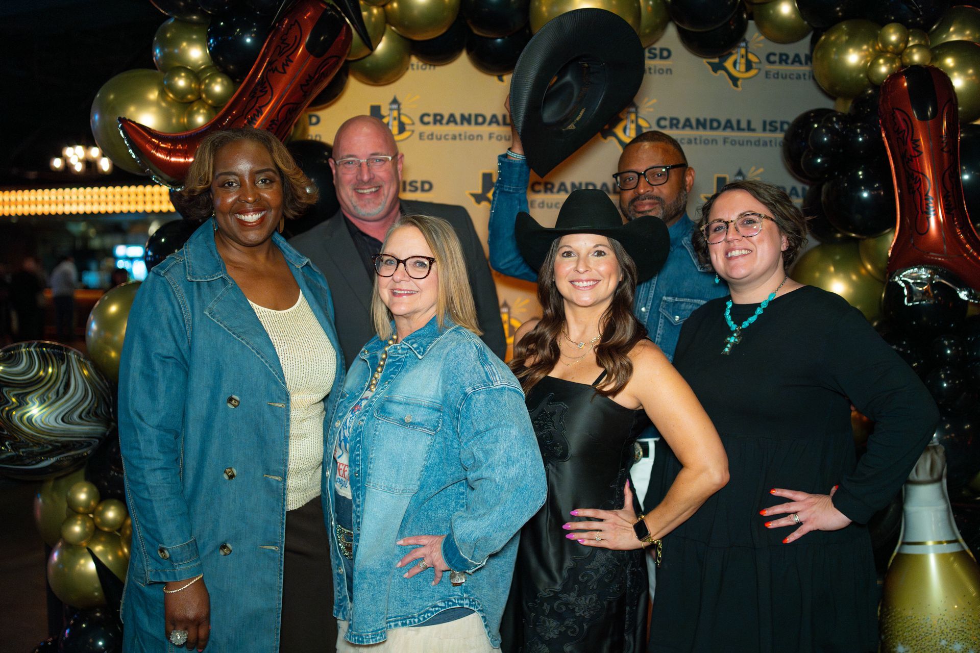 Group of six people posing at a Western-themed event. Some wear cowboy hats, smiles, and a backdrop of balloons.