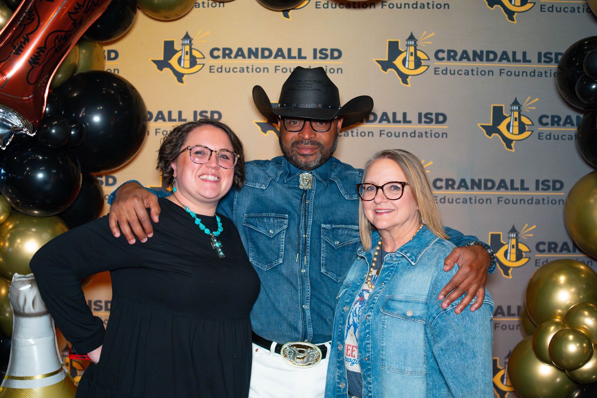 Three people posing at an event with black and gold balloons. The man in the middle wears a cowboy hat and jean jacket.