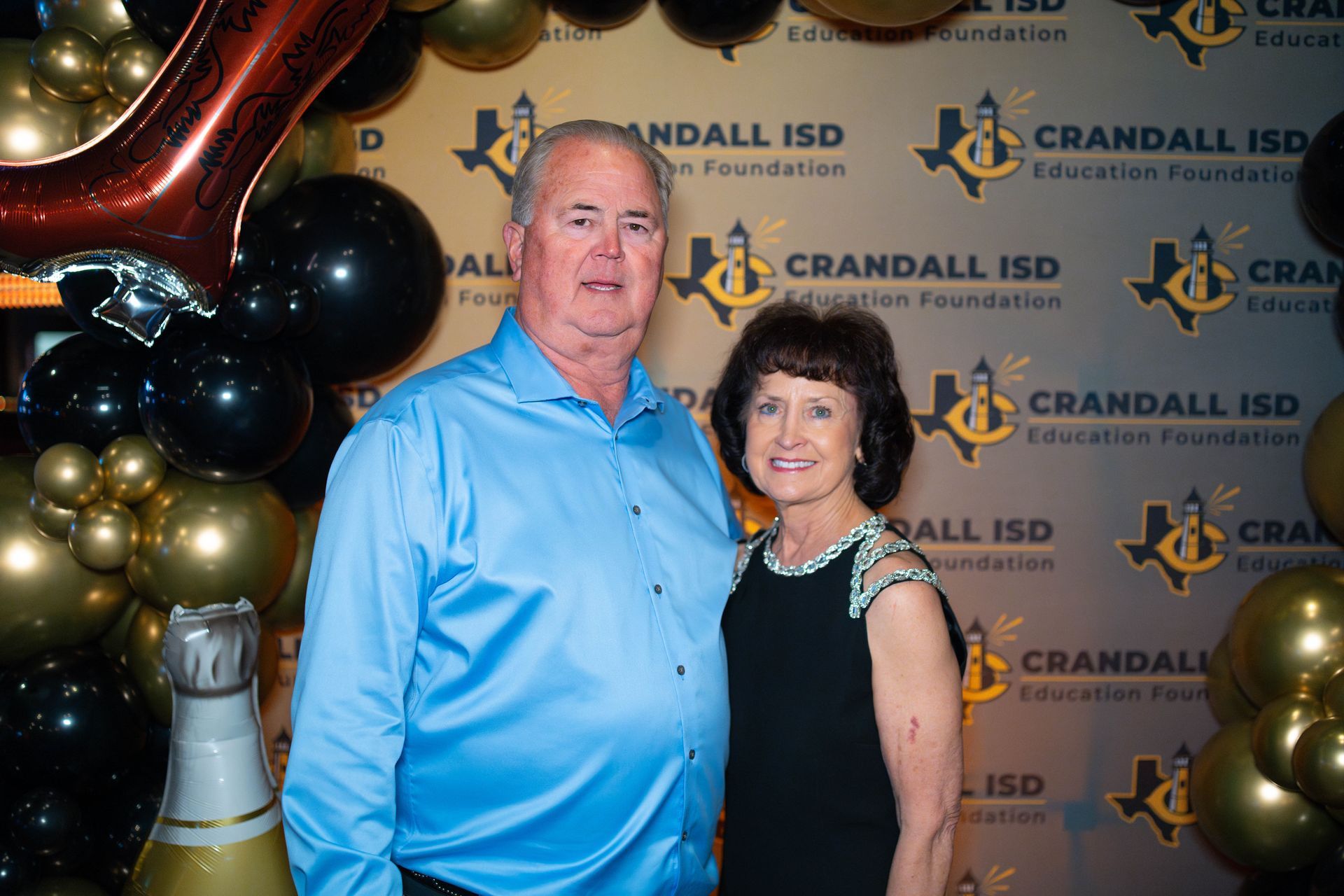 A man and woman smiling, posing in front of a Crandall ISD Education Foundation backdrop with black and gold balloons.