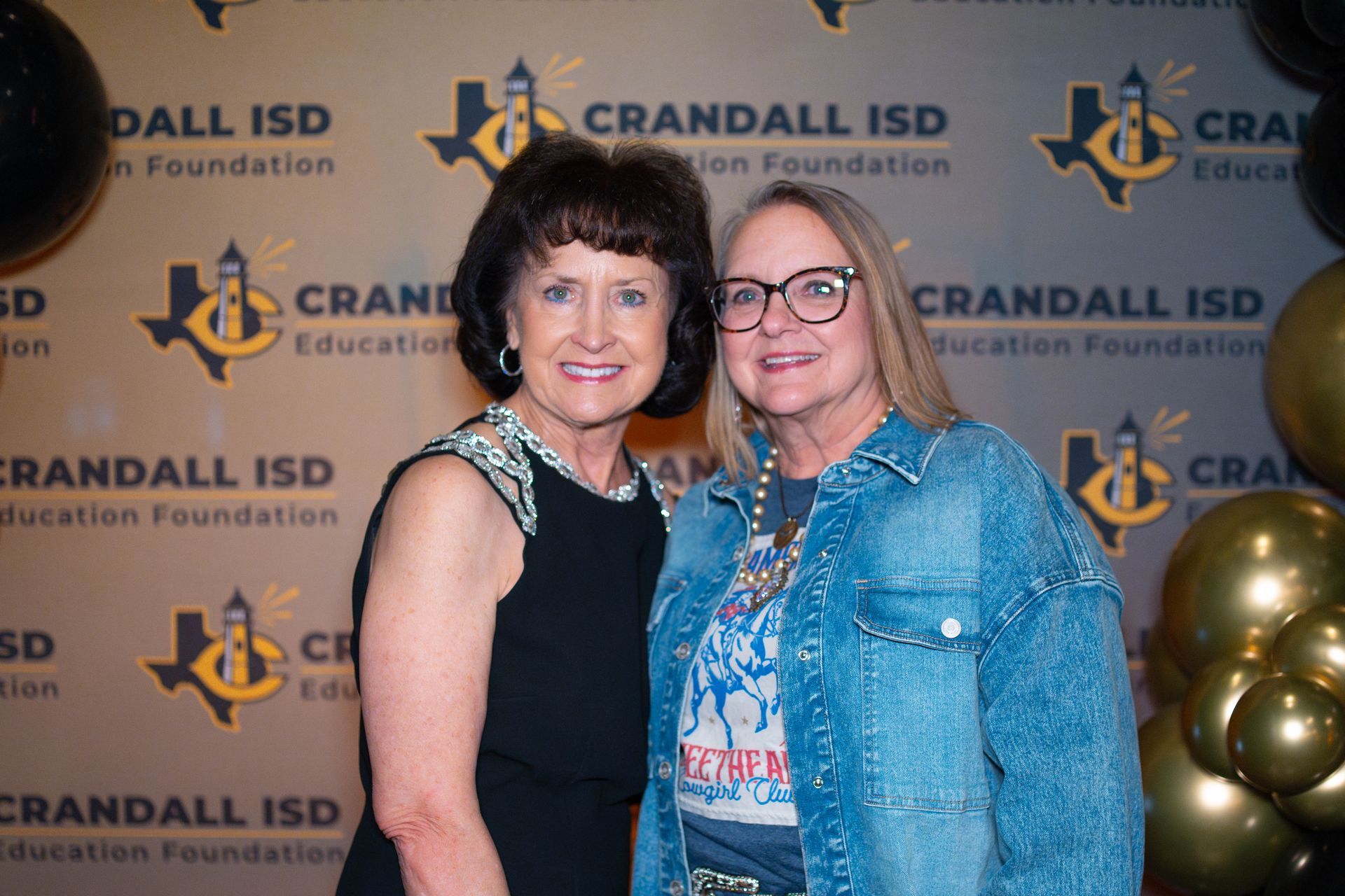 Two women smiling at a Crandall ISD Education Foundation event; a black and gold backdrop.
