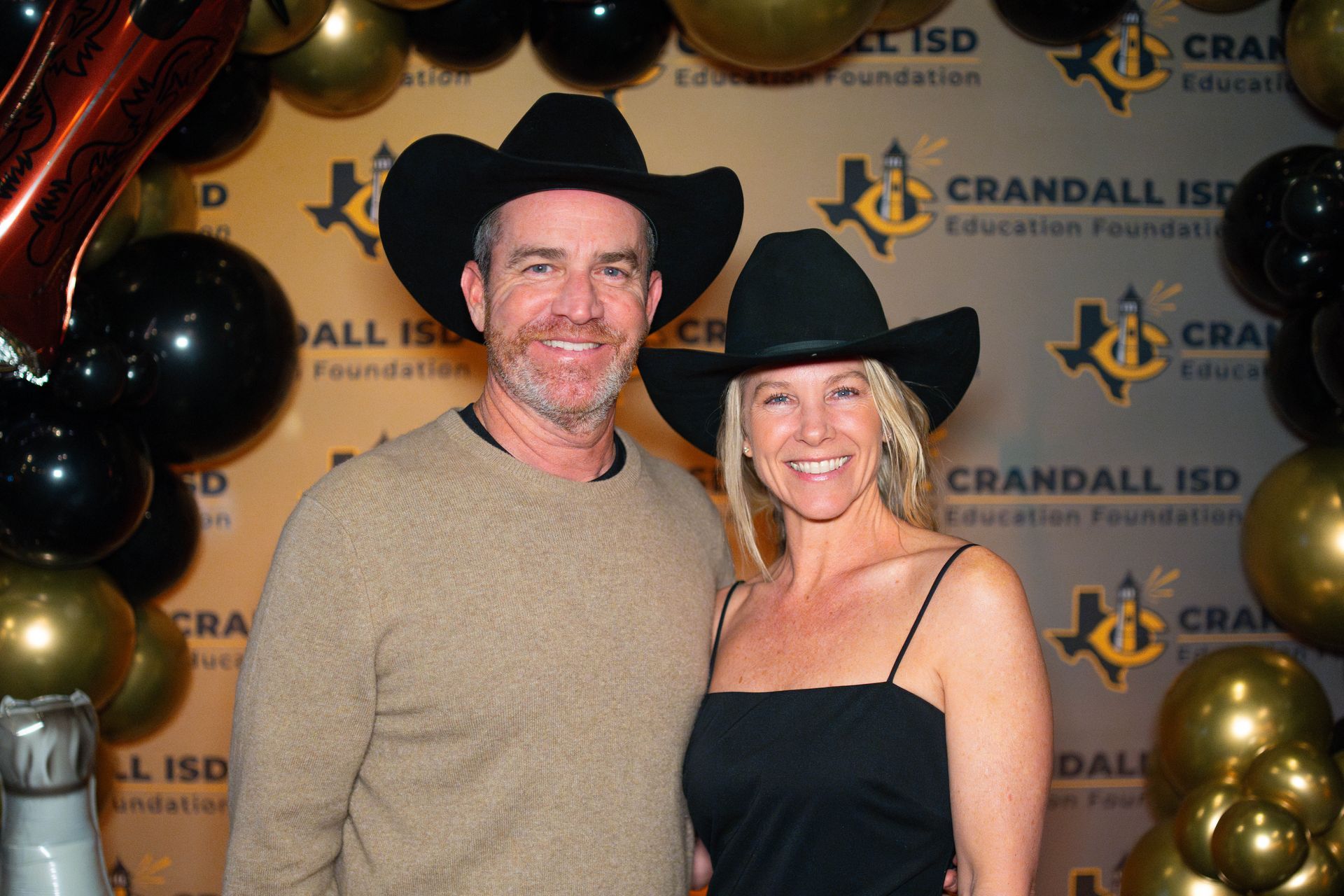 Man and woman in cowboy hats pose at a Crandall ISD Education Foundation event, smiling in front of gold and black balloons.