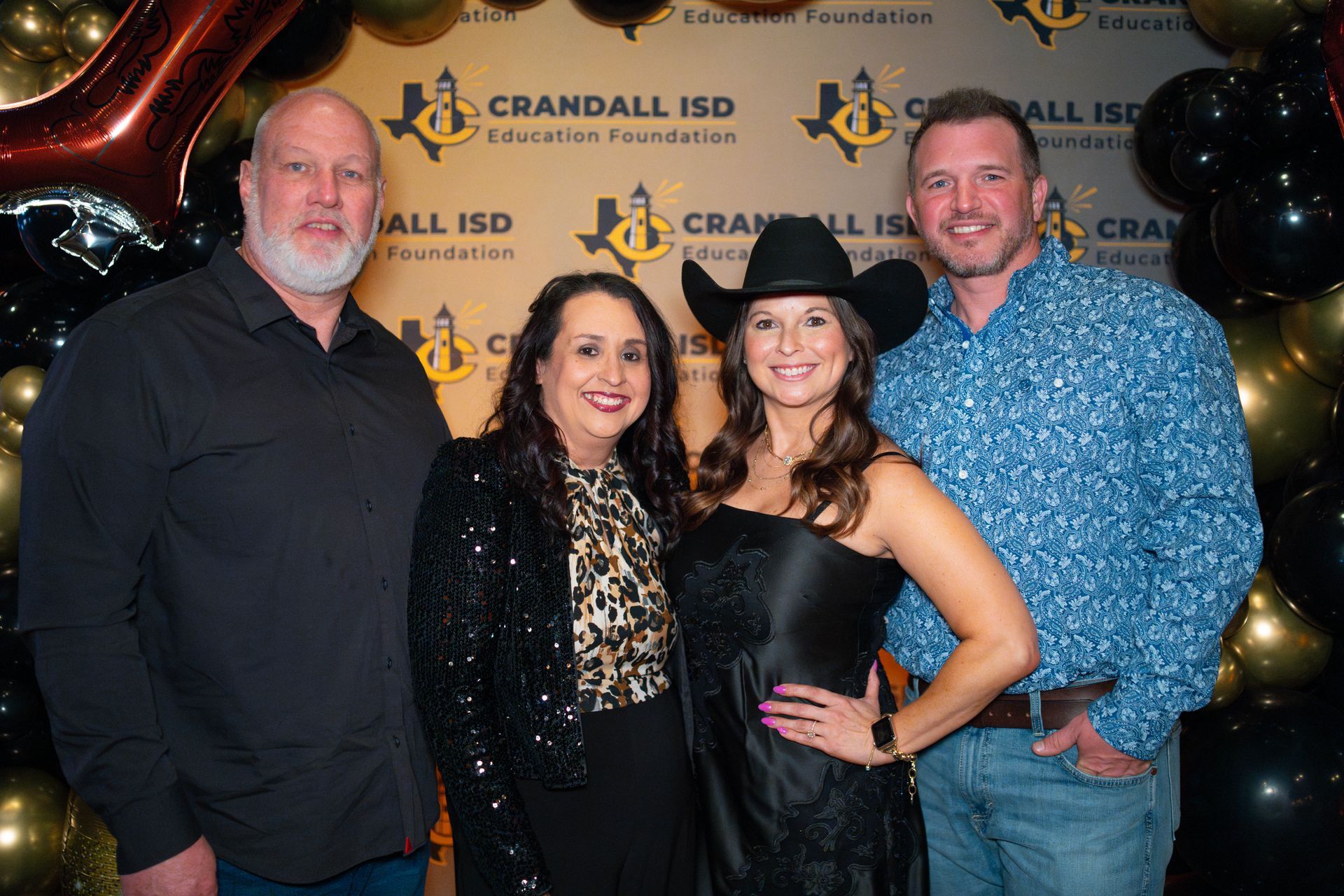 Four people pose for a photo at an event, smiling. Two women, a man in a cowboy hat, and a man in a patterned shirt stand near a backdrop with balloons.