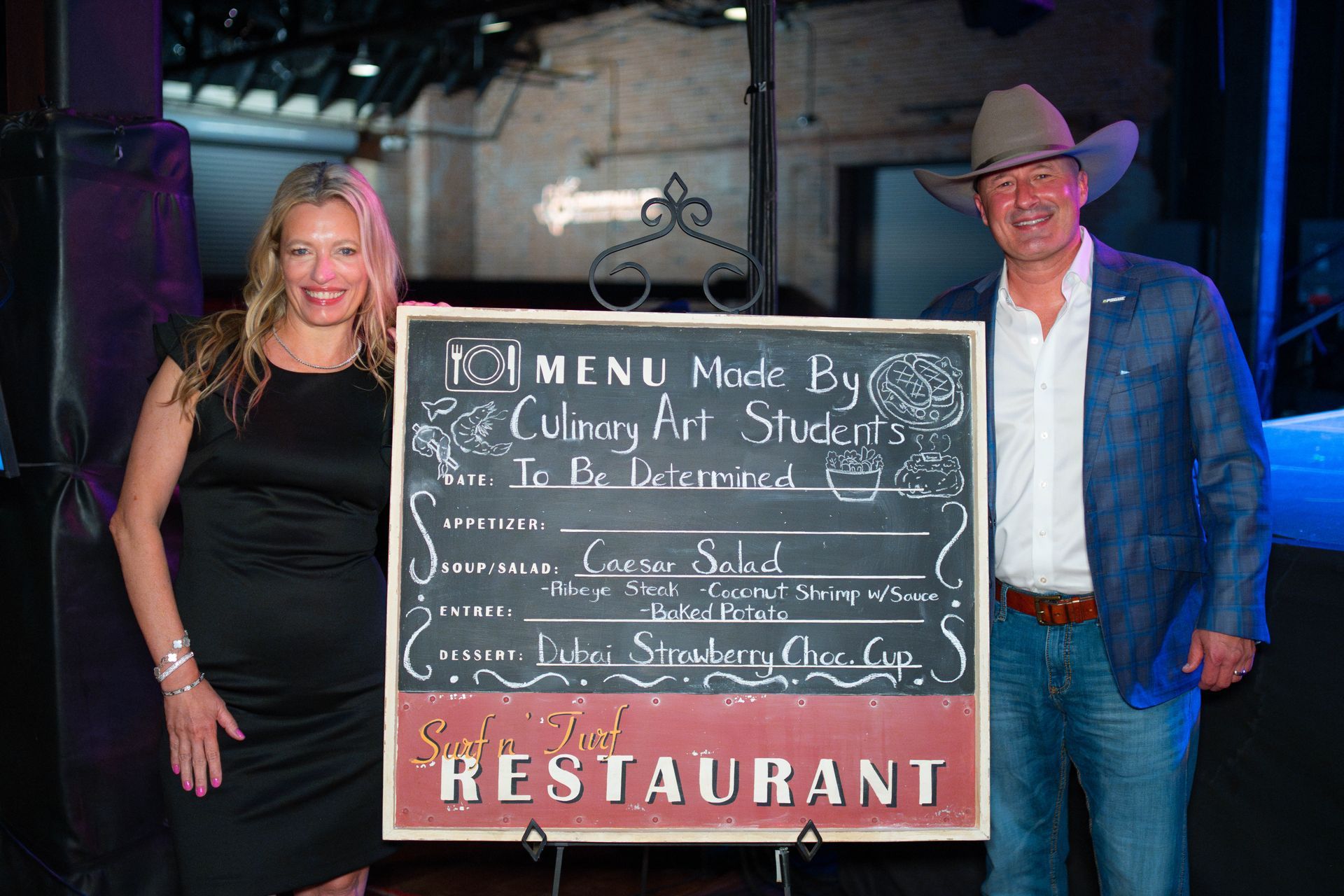 Woman and man holding chalkboard menu; restaurant setting. Menu lists items: Corn Salad, Rib Eye, Strawberry Cheese.
