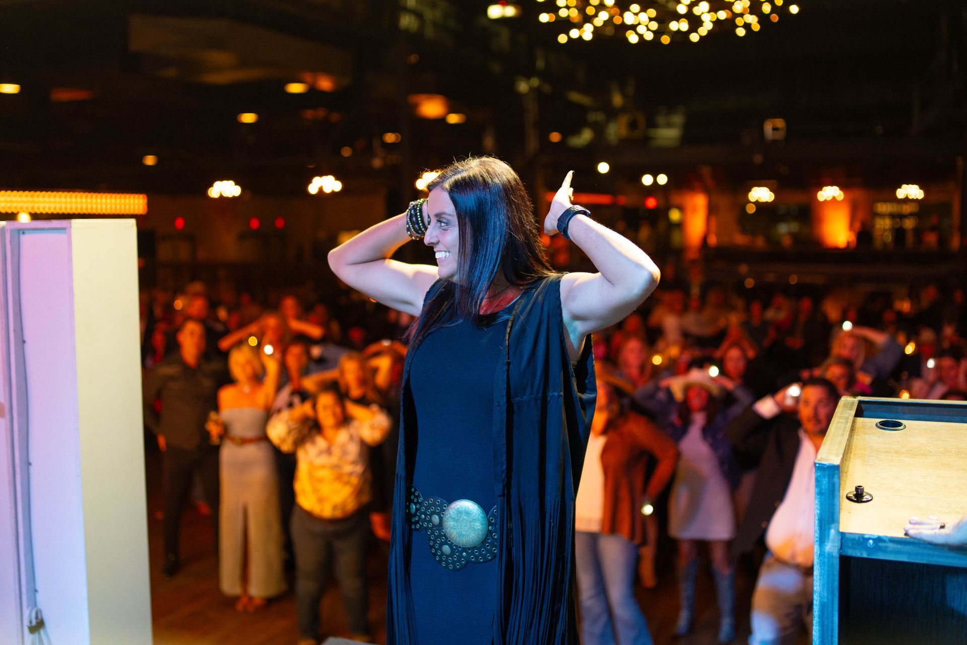Woman on stage with hands up, facing crowd. Dark dress, stage lights, large audience.