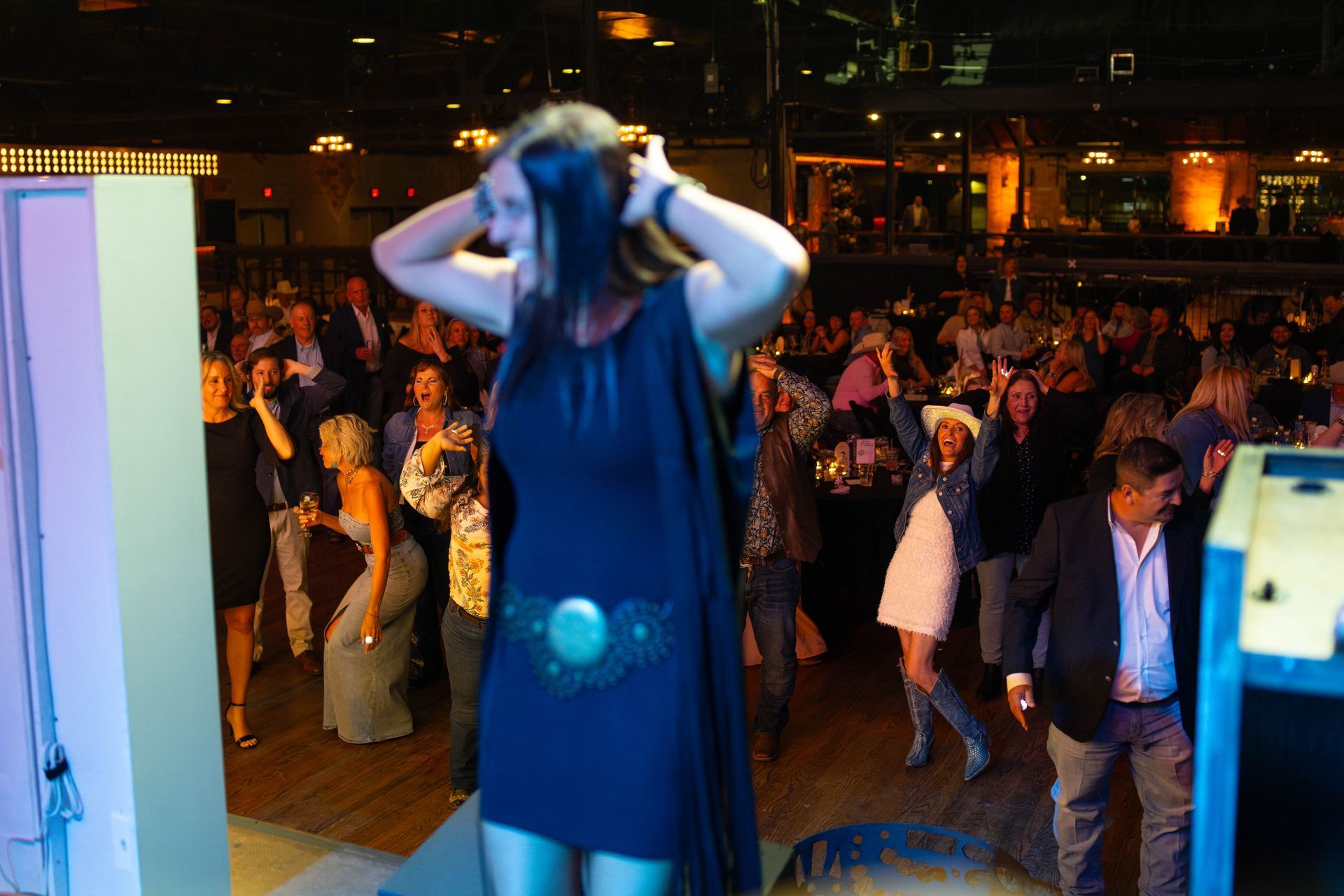 Woman in black dress with arms raised, performing on stage in front of a dancing crowd.