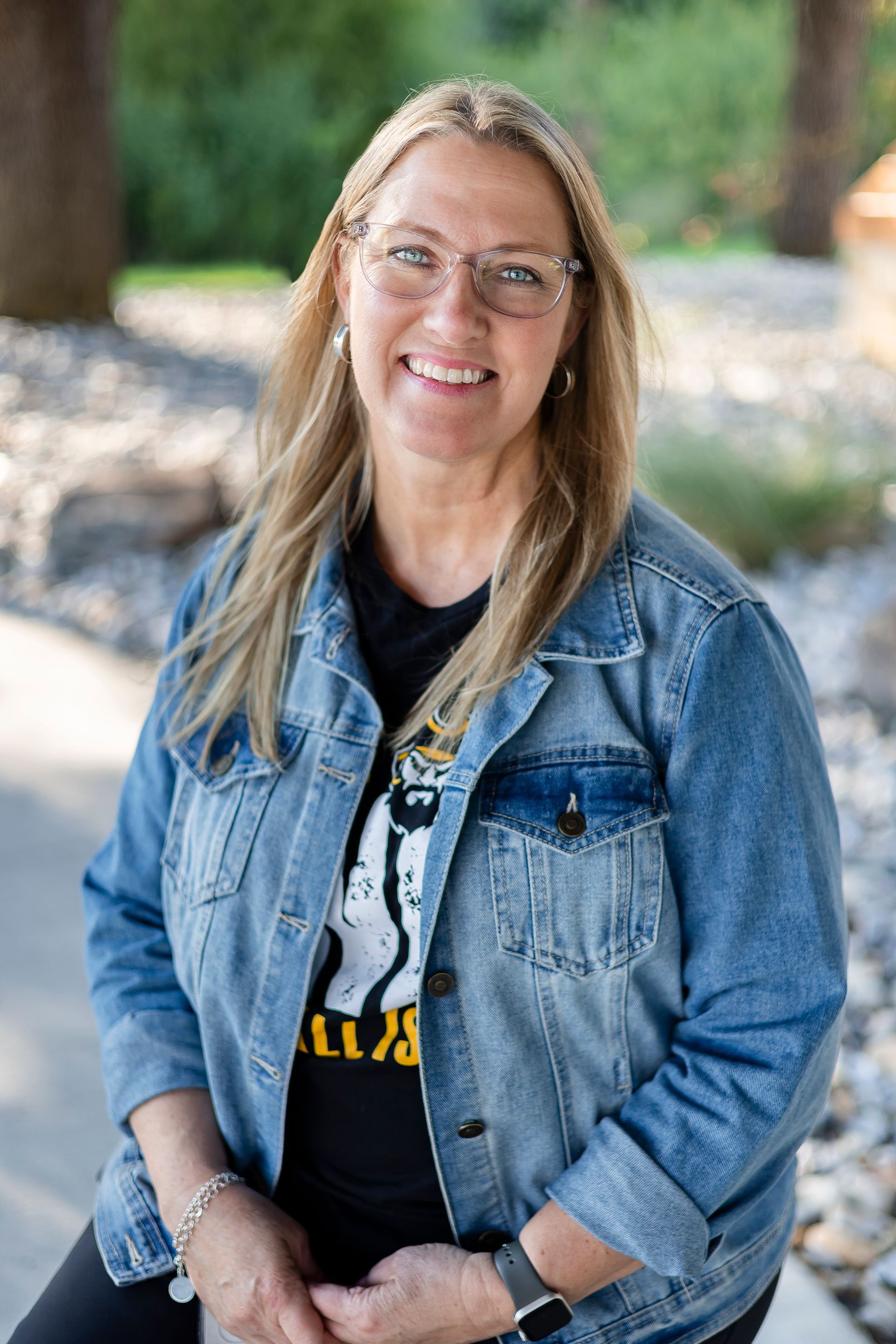 Woman with blonde hair, glasses, smiling, wearing a denim jacket and black shirt, outside.