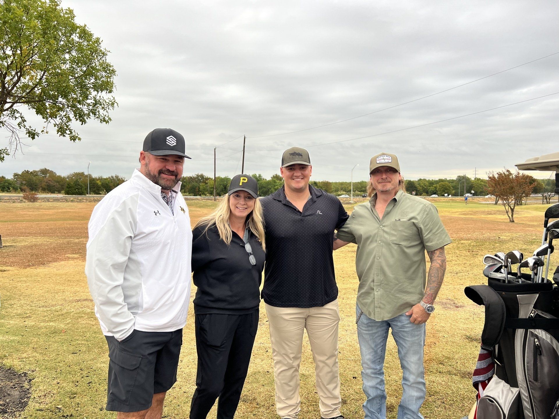 Four people smiling, posing outdoors near golf course; cloudy day.