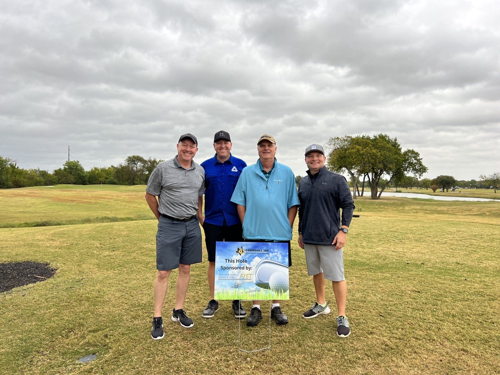 Four men standing on a golf course, holding a sign after a tournament. Overcast day.