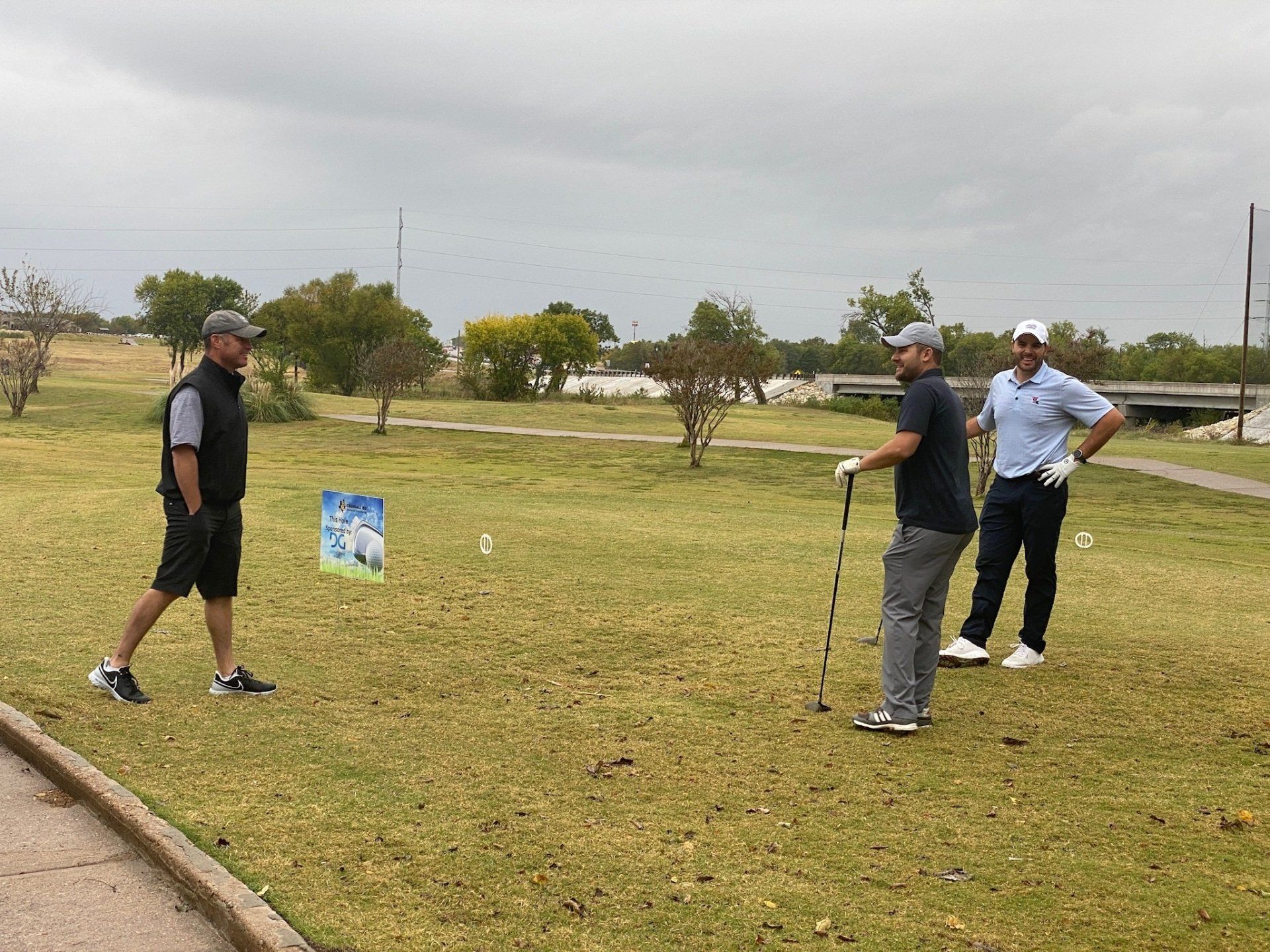 Three men on a golf course. One holds a club, two are watching. Overcast day.