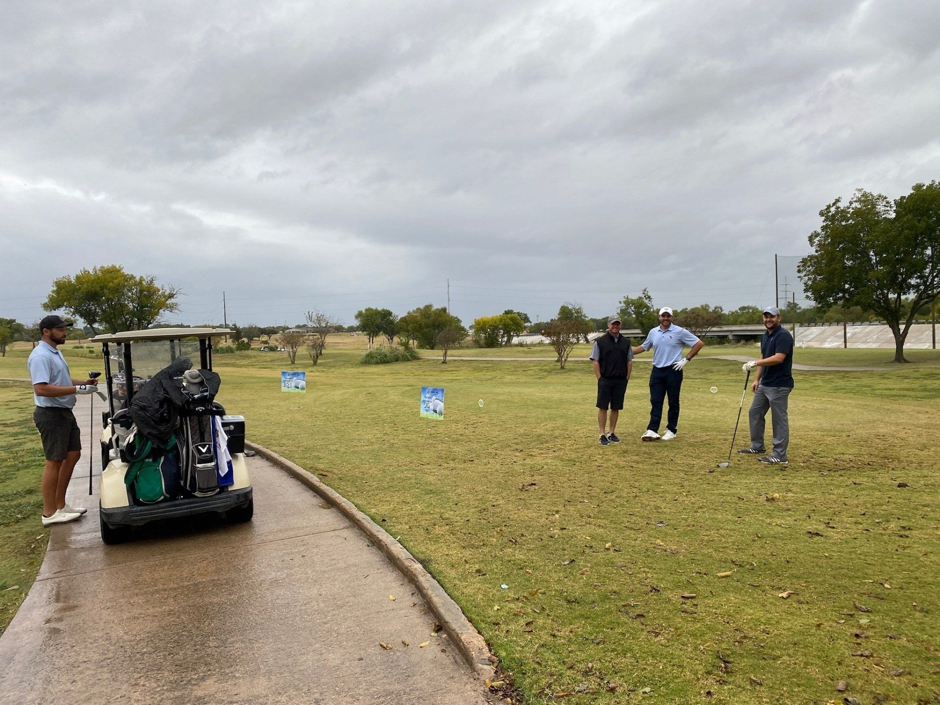 Four men on a golf course, one with a golf cart. Overcast sky, green grass, and trees.