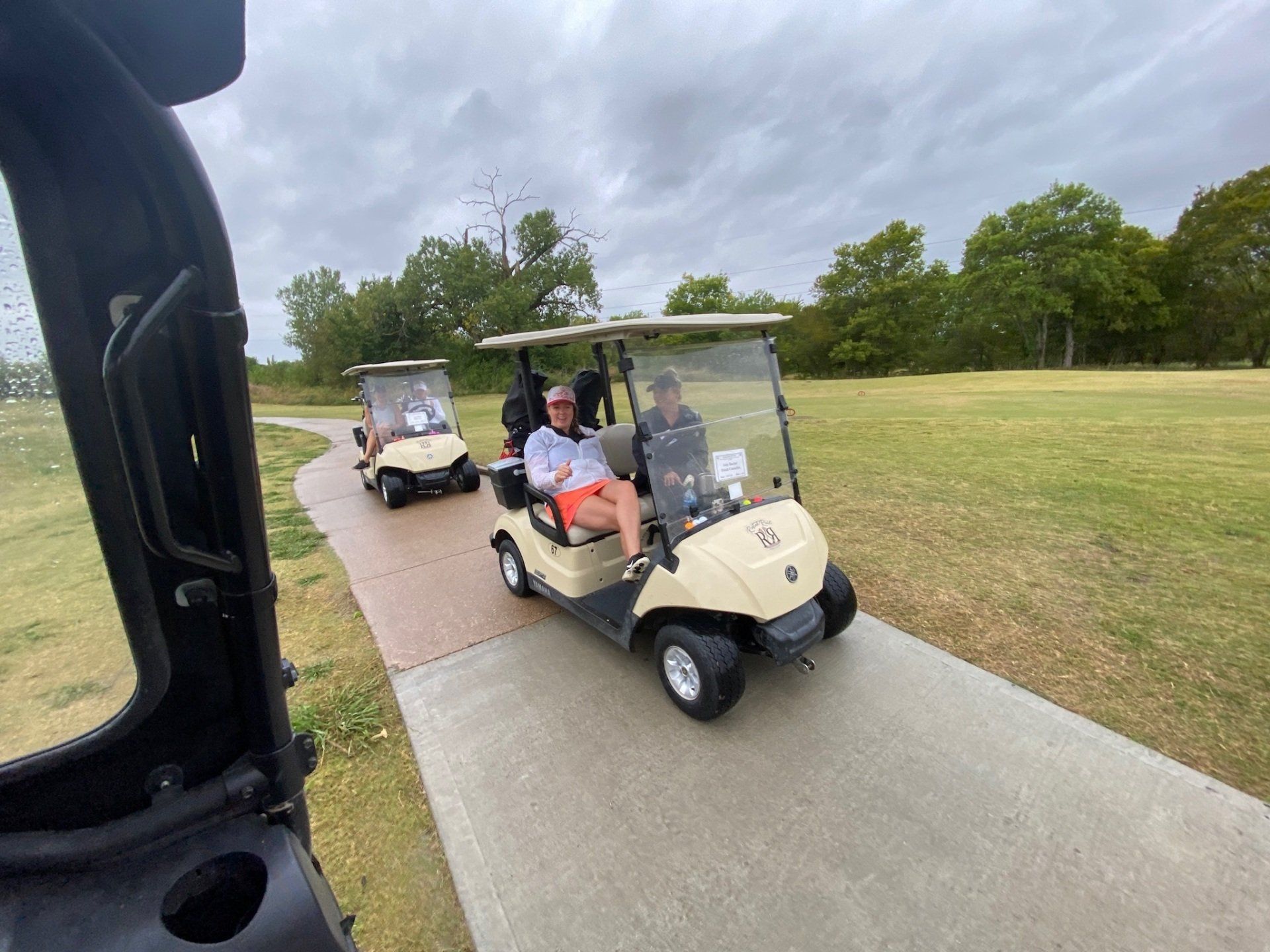 Golfers in beige golf carts on a paved path on a cloudy day at a golf course.