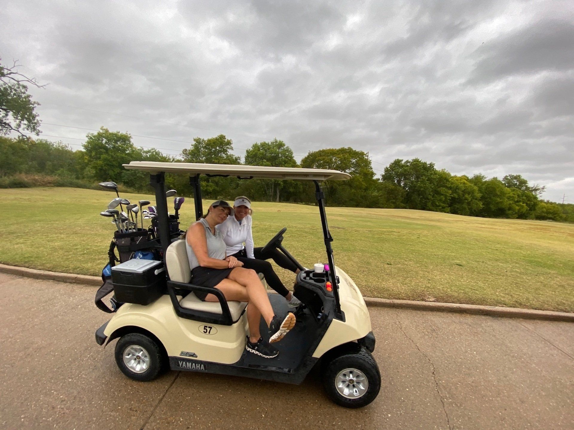 Two women in a golf cart on a course, cloudy day. One is driving; the other is resting.