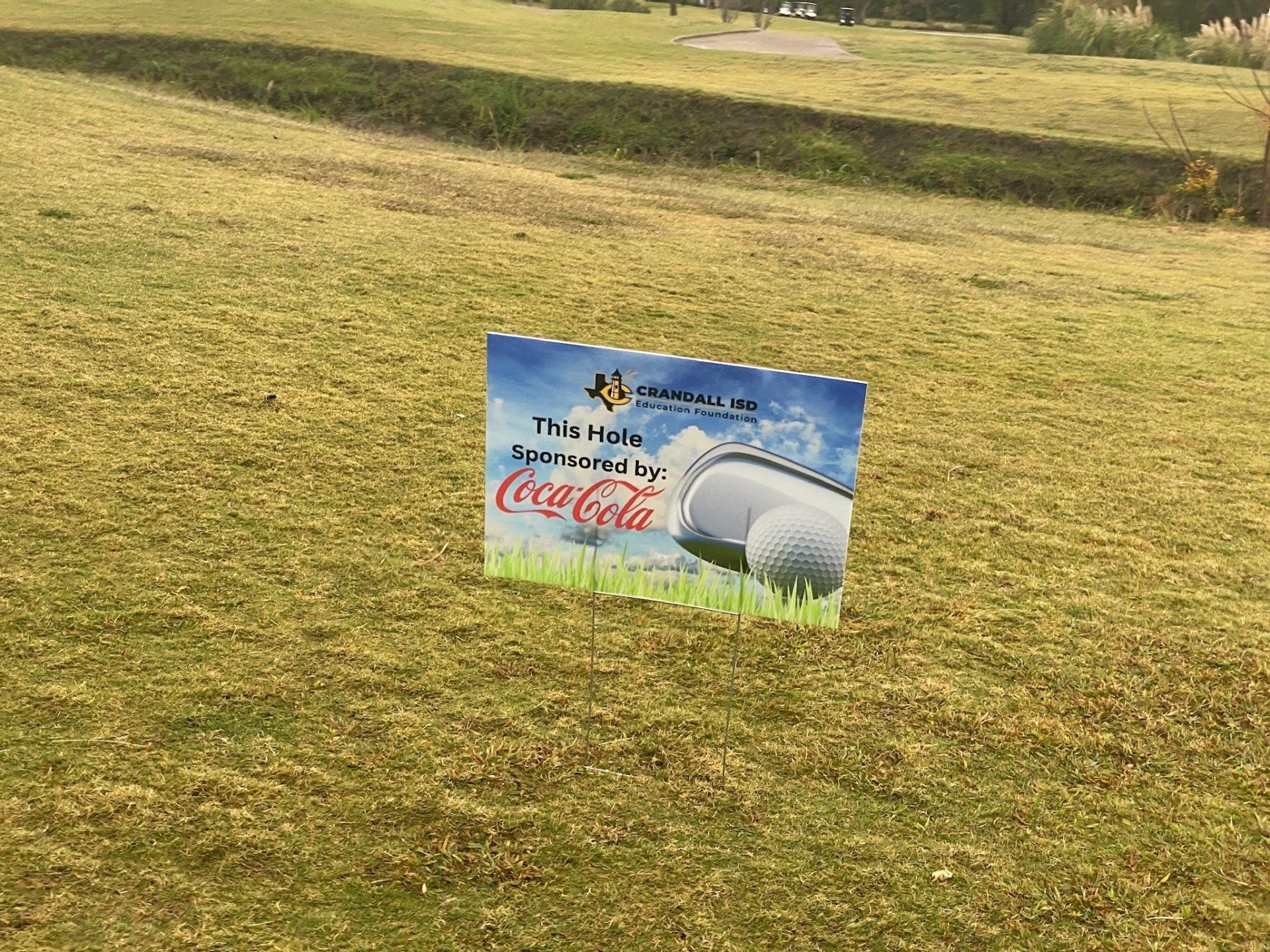 Sign on a golf course advertising Coca-Cola, with a golf ball graphic and text: 