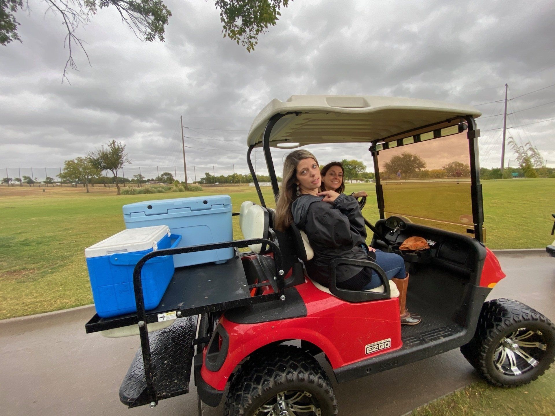 Two women in a red golf cart, smiling, with a blue cooler on the back, cloudy outdoor setting.