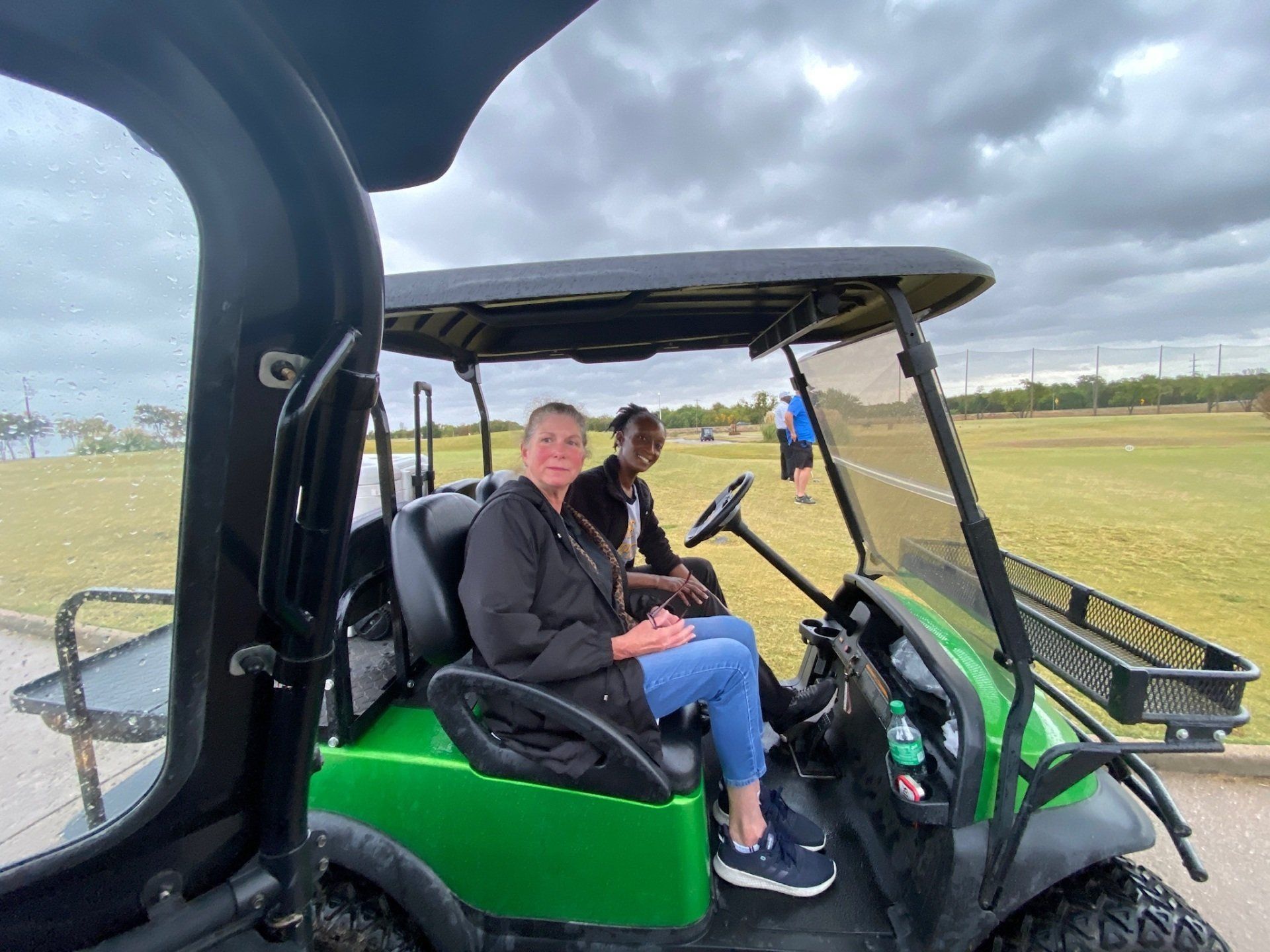 Two people in a green golf cart on a course. Overcast day, woman smiling, black man in the back.