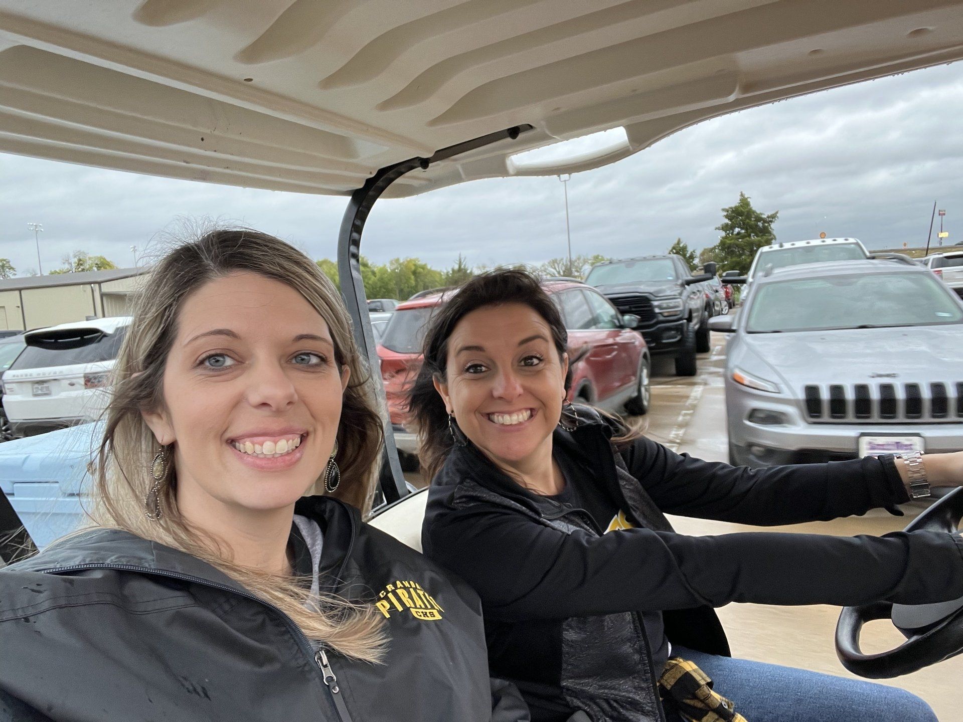 Two women smiling in a golf cart outdoors; parked cars visible.
