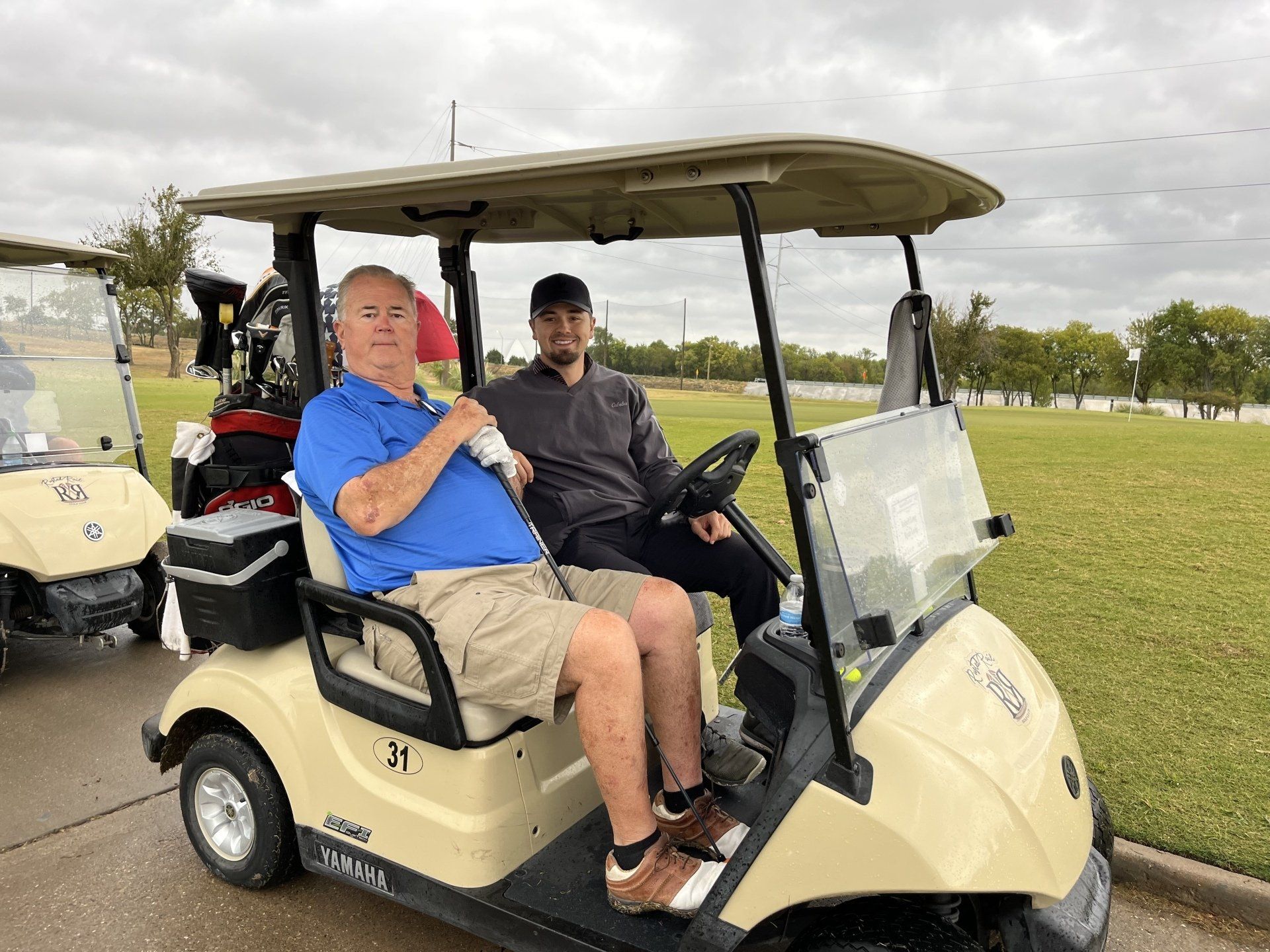 Two men in a golf cart, outdoors on a golf course on a cloudy day.