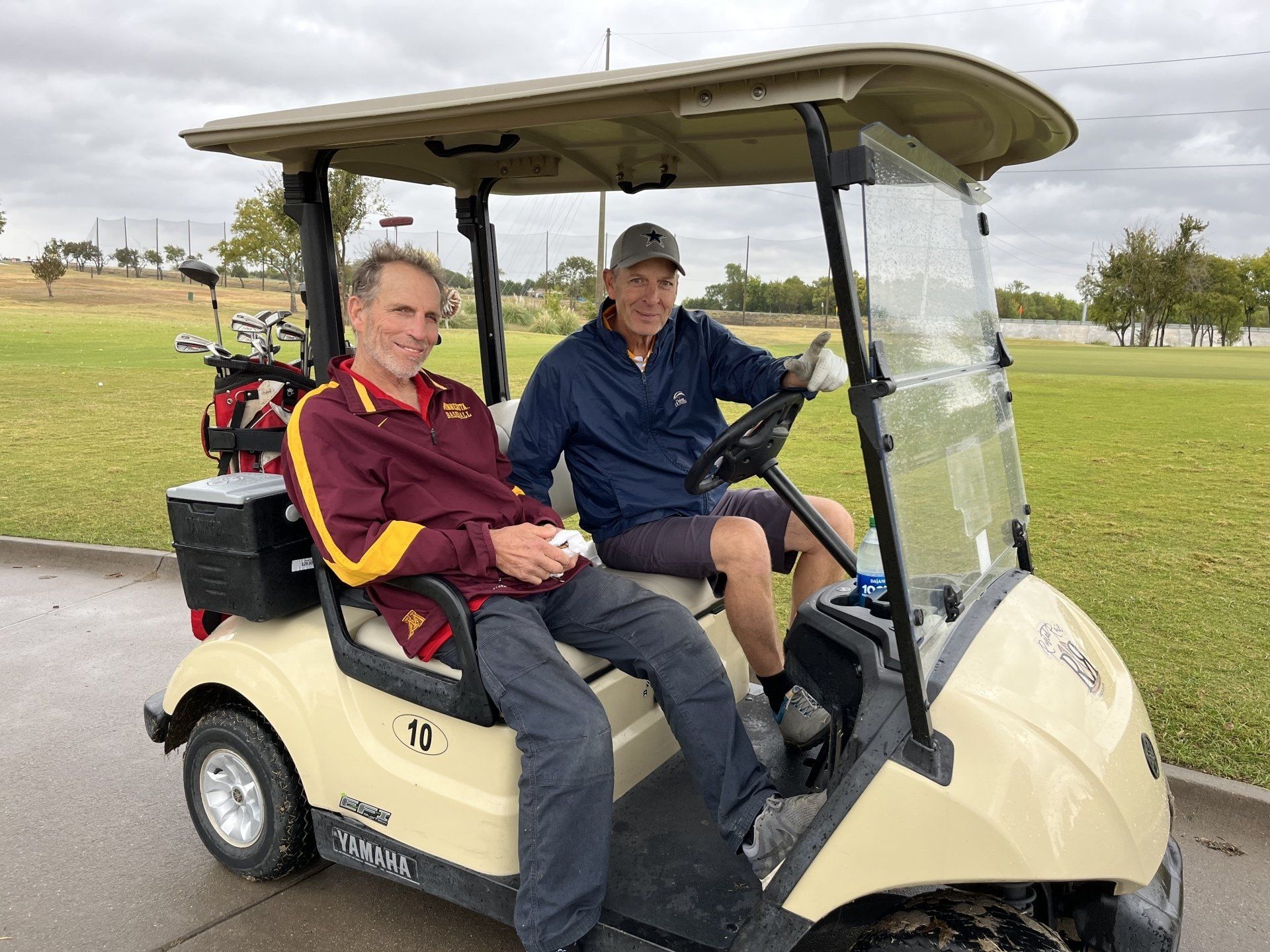 Two men in a golf cart on a golf course, one driving, the other smiling, overcast day.