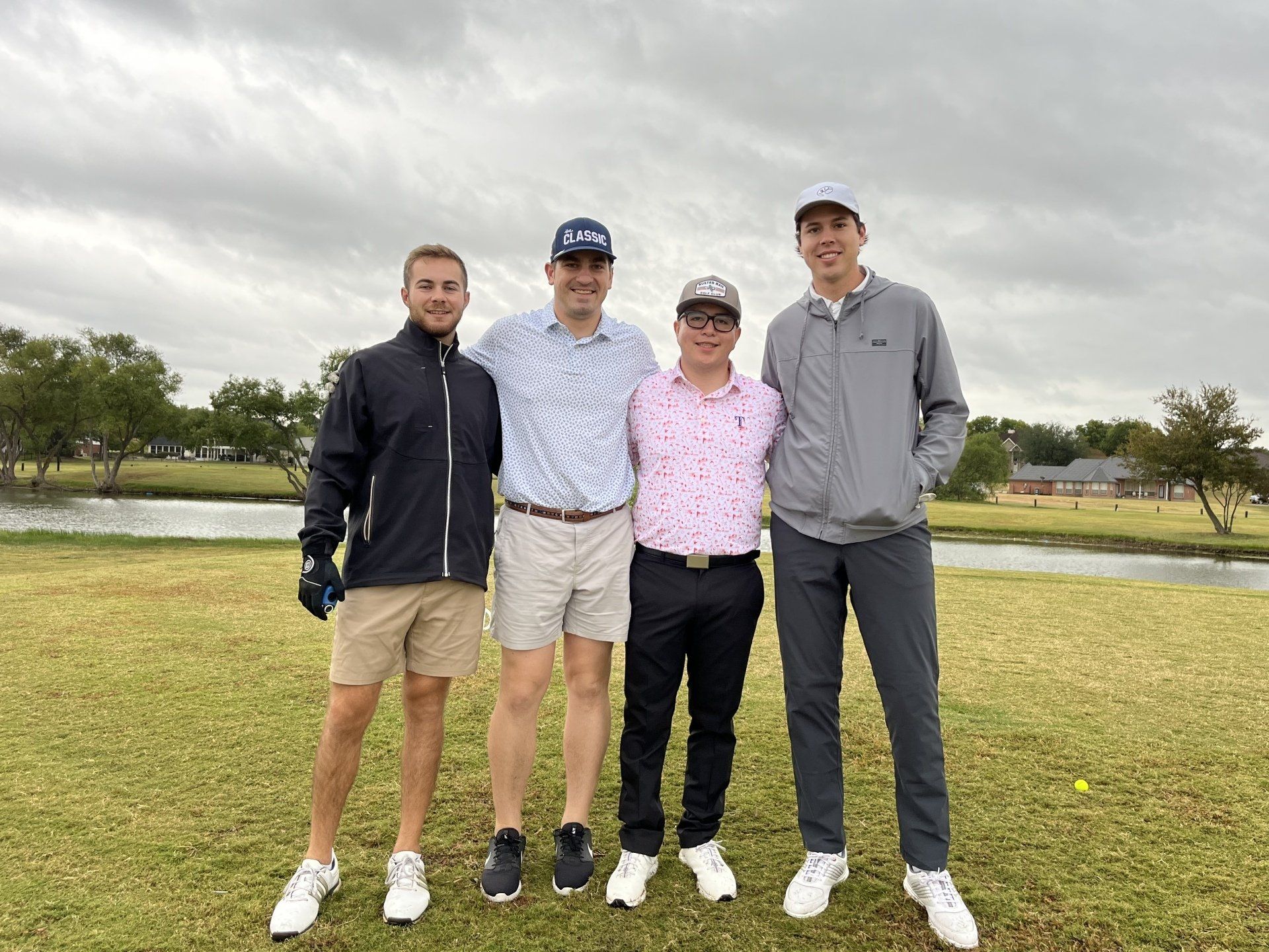 Four men in golf attire stand on a green with a pond and cloudy sky; posing after a game.