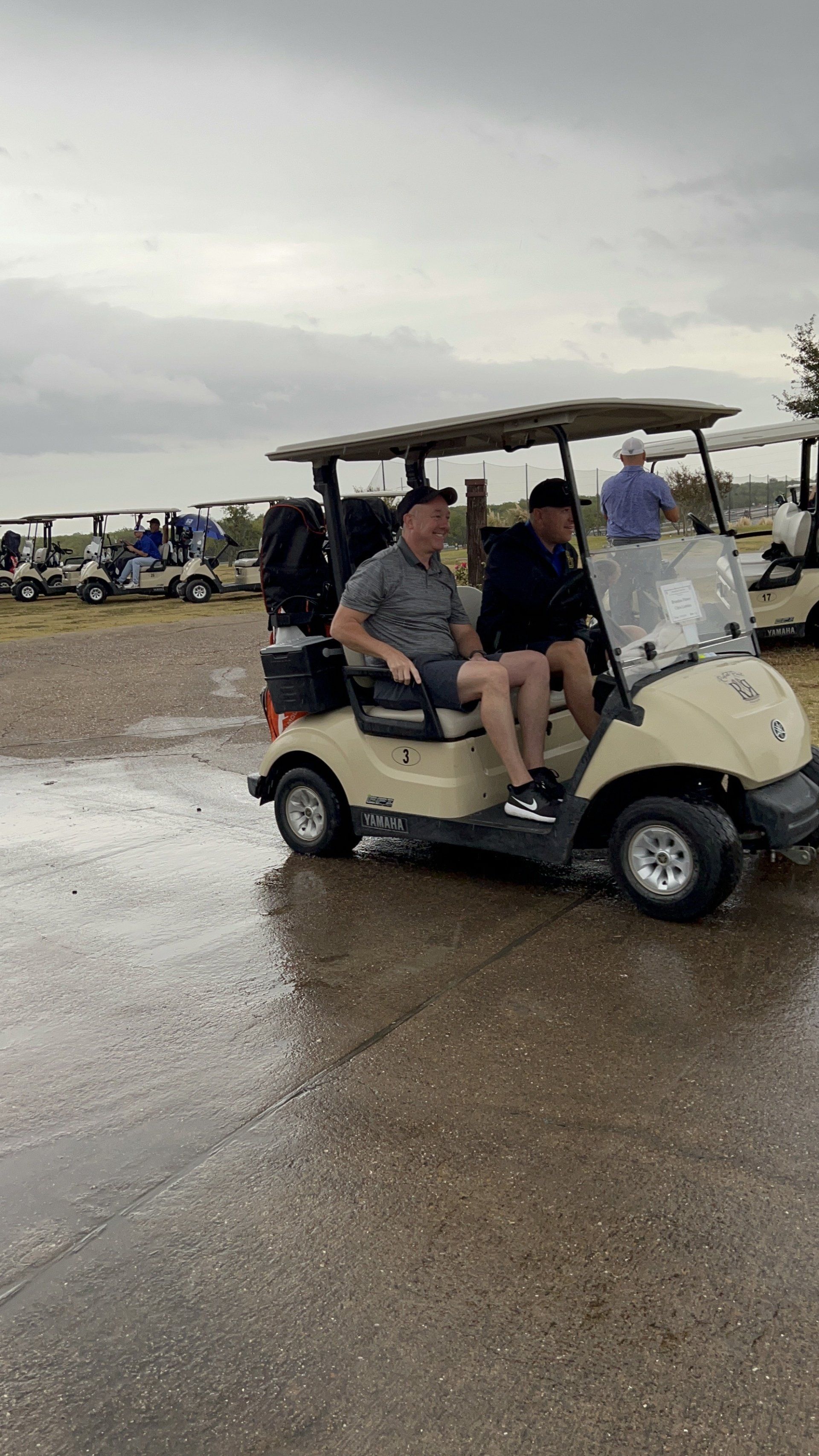 Two men in a tan golf cart on a wet course, with other carts in the background under a cloudy sky.