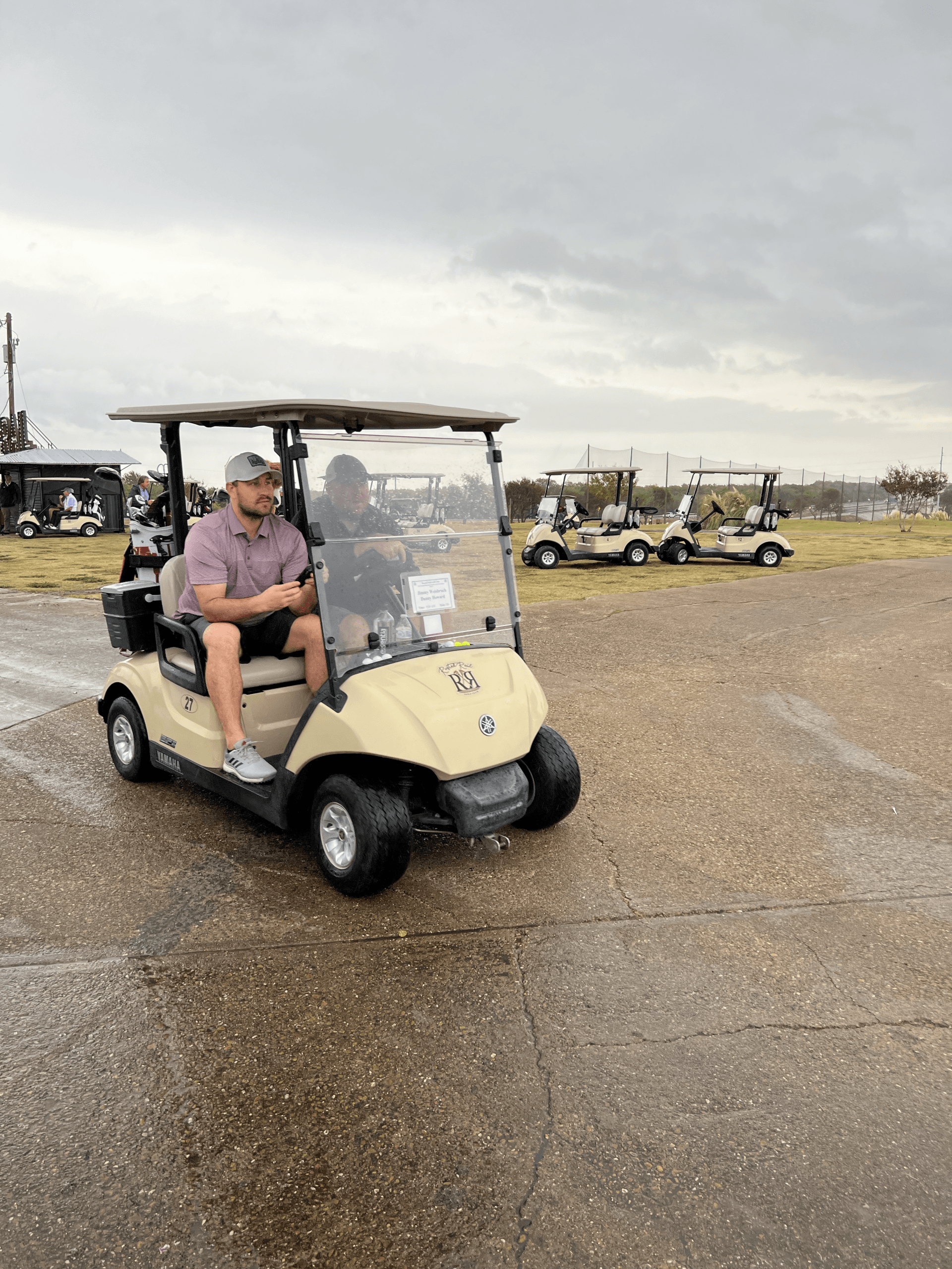 Man in a beige golf cart on a wet lot. Other golf carts in background under overcast sky.
