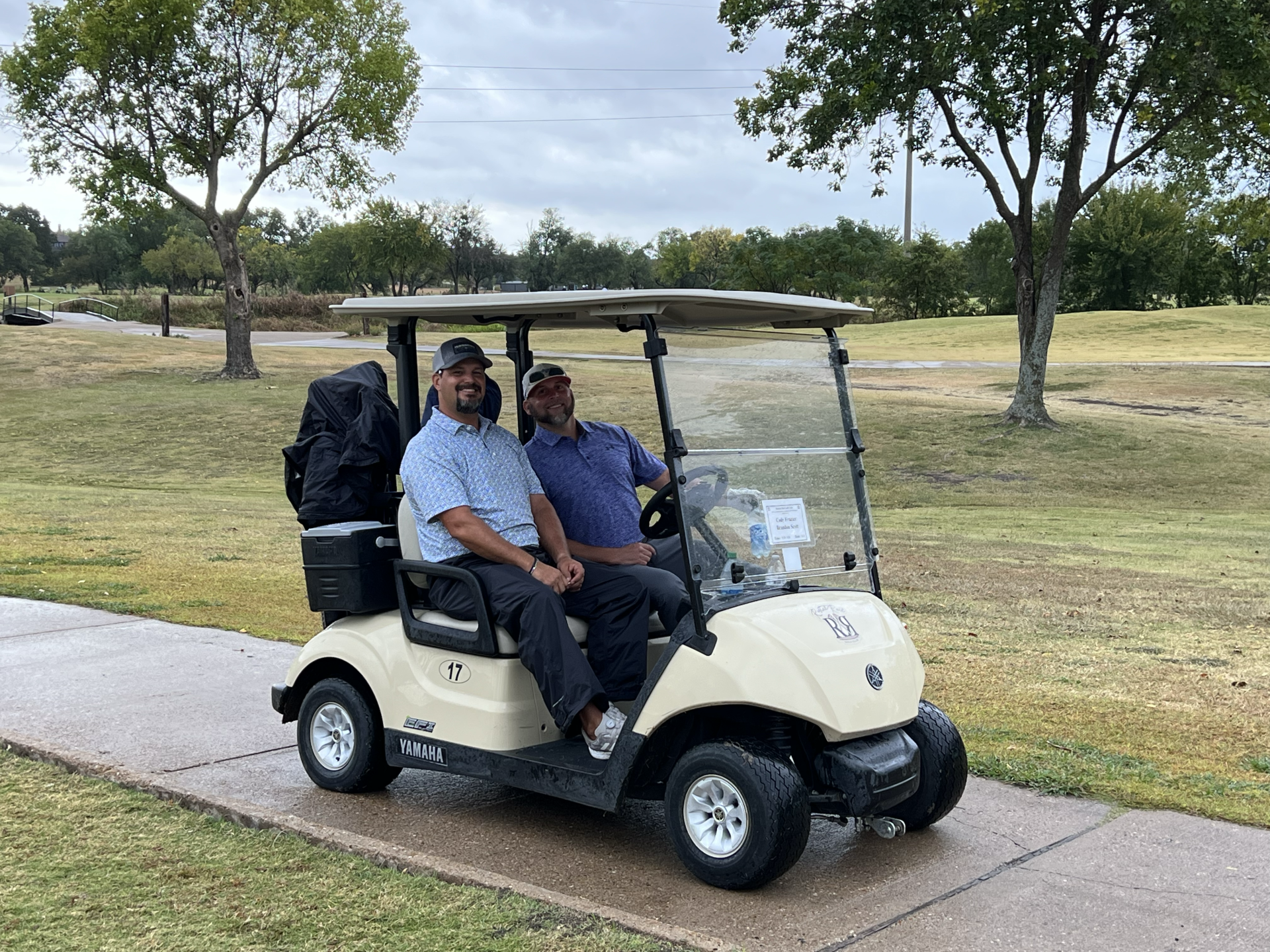 Two men smiling in a beige golf cart on a paved path at a golf course.
