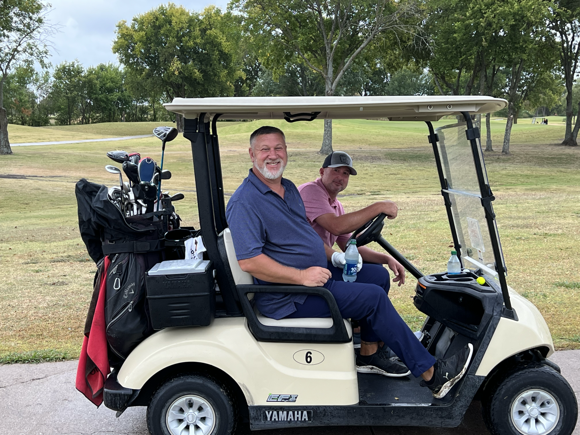 Two men in a beige golf cart on a course. One smiles while the other drives, trees in background.