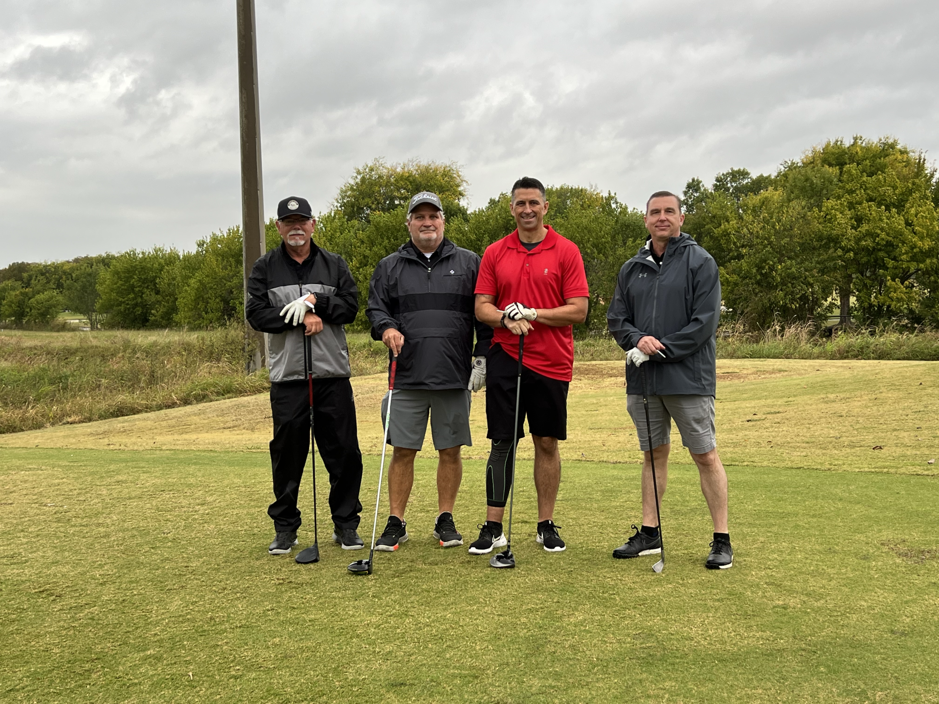 Four men stand on a golf course, each holding a club. Overcast sky, green grass.