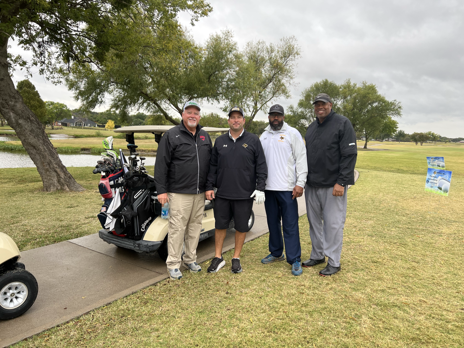 Four men stand next to a golf cart on a green course. Cloudy day, lake in the background.