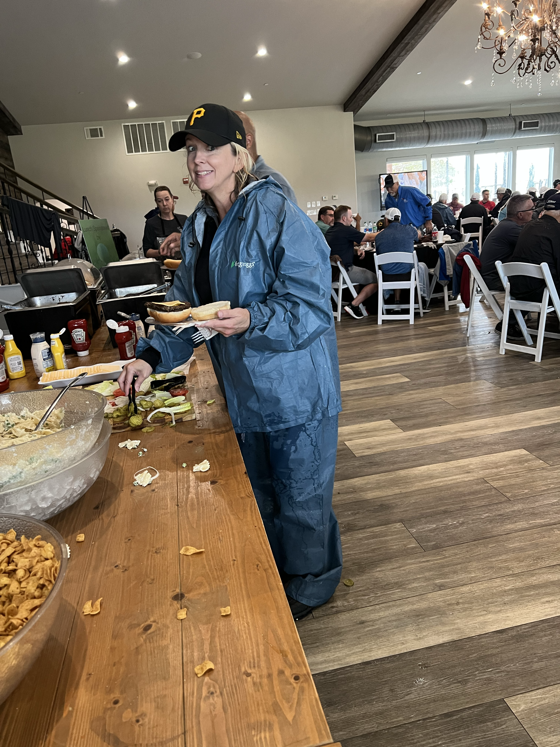 Woman in blue jacket and hat serving food at a buffet, other people in background.